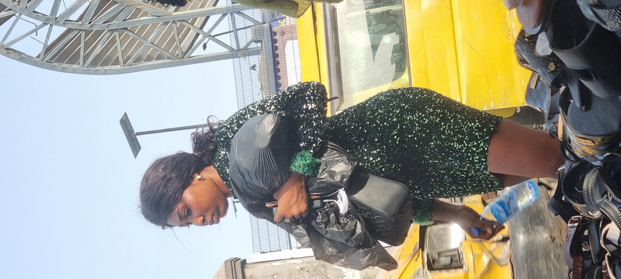 A woman stands in what appears to be a market or bustling street environment in Lagos, Nigeria. She is wearing a green, sequined dress, and carries a black handbag and a black plastic bag containing items that may include a green decorative element. She also holds a water bottle. The background features a yellow vehicle, possibly a bus or taxi, and an assortment of footwear is displayed for sale. Above, there is a metal awning structure, indicating an outdoor location under a shelter.