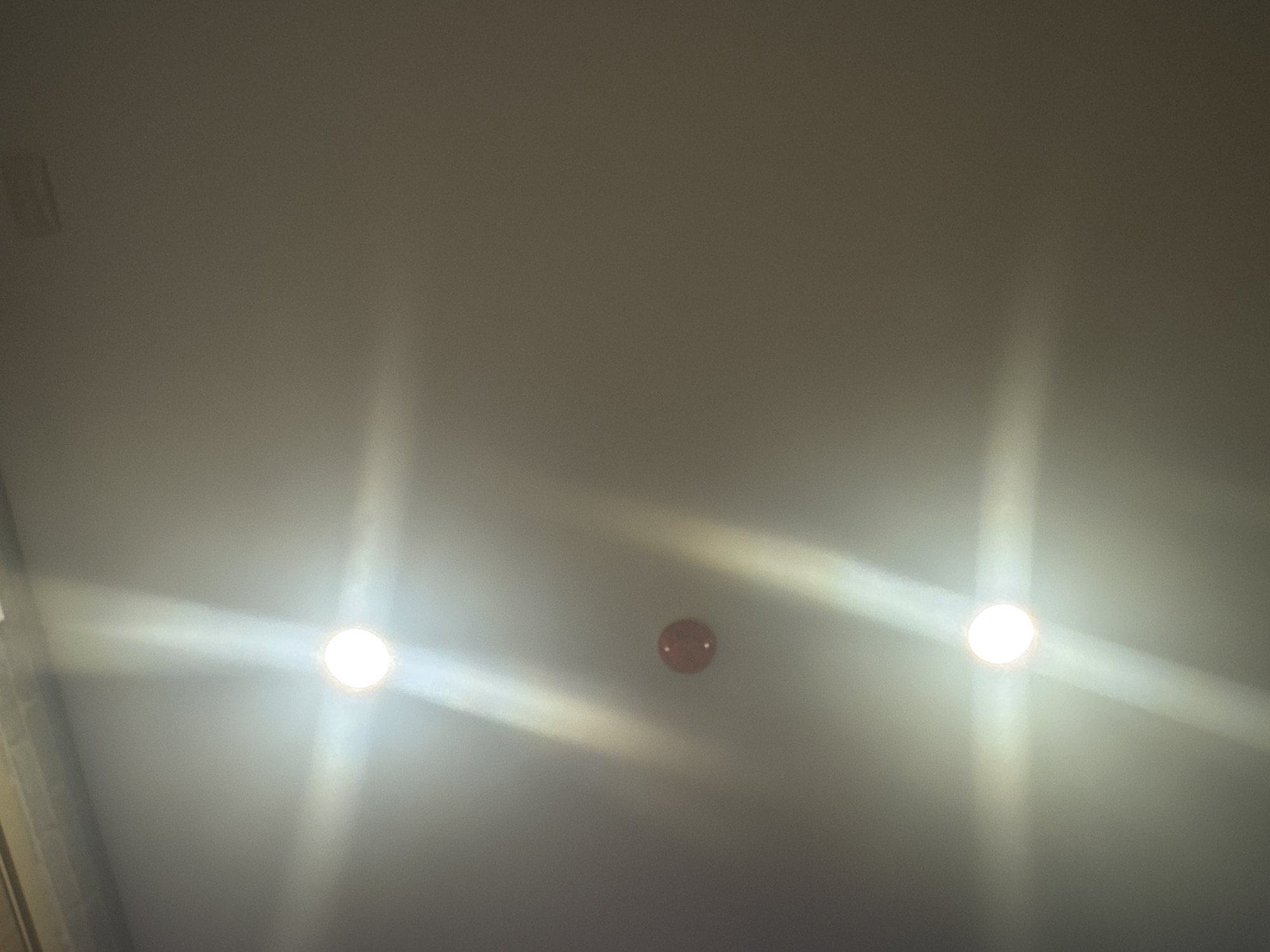 This is a photograph of a ceiling in an indoor environment. Two circular lights are visible, one above the other. The top light is bright and appears to be a recessed ceiling light, casting a strong glow and lens flare. Below it, slightly to the left, is a small, round, red object, possibly a smoke detector or a camera. The bottom light is also a bright, circular recessed light, similar to the top one, and it also emits a strong glow and lens flare. The ceiling appears to be a plain, light-colored surface, possibly painted drywall or plaster, with some subtle texture. There are no people or any other discernible objects in the frame. The lighting conditions suggest a low-light environment where artificial lighting is the primary source of illumination. The image does not contain any identifiable features that would allow for a specific location within Bangkok, Thailand, to be determined.