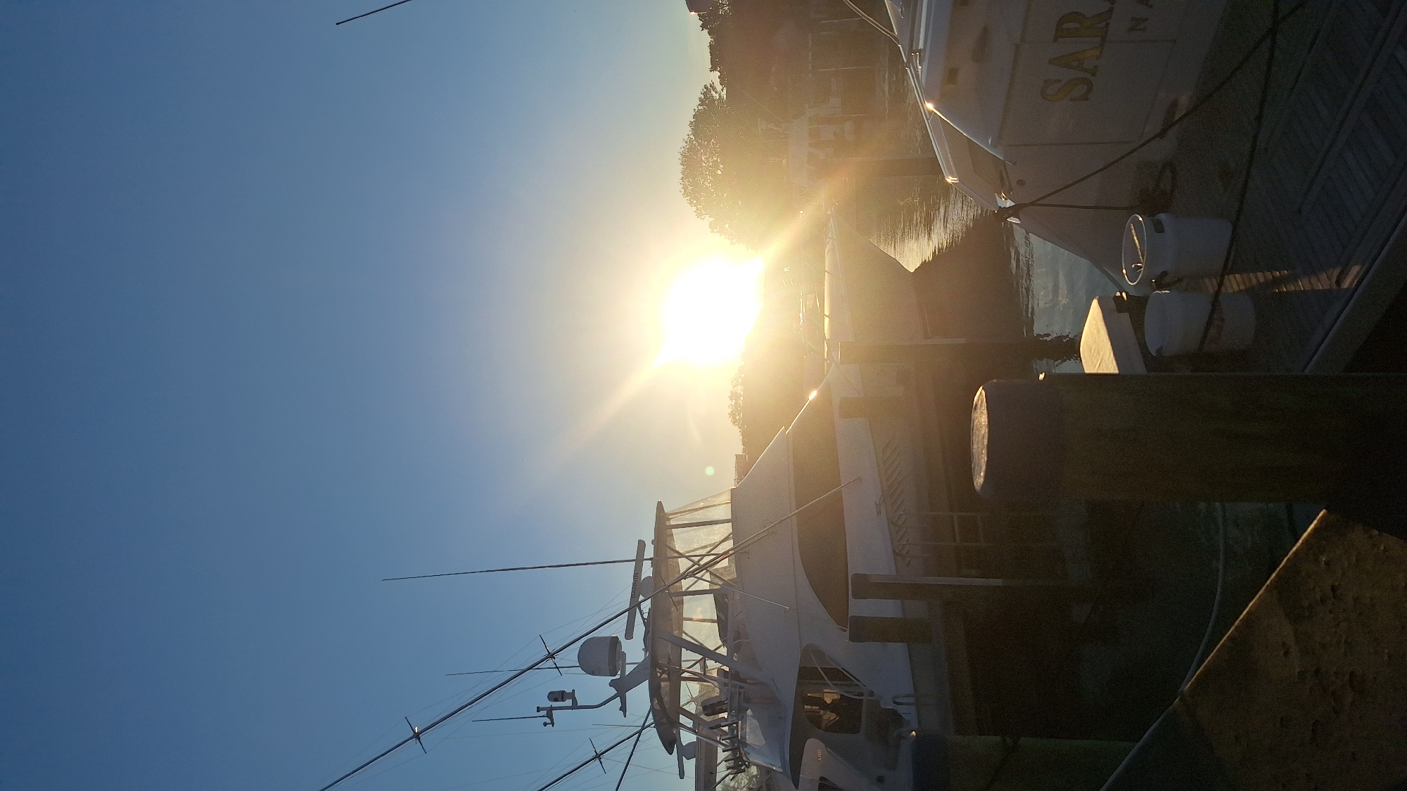 The image depicts a serene harbor scene in Nantucket, Massachusetts, USA, illuminated by the warm glow of a setting sun. In the foreground, sturdy wooden pilings anchor a dock. Moored boats, including a large white yacht with fishing poles prominently displayed, occupy the middle ground. The yacht's name, "SARAH," is partially visible in golden lettering on its stern. The water reflects the sky and the boats, creating a tranquil atmosphere. Trees and a building line the distant shore, silhouetted against the vibrant orange and yellow hues of the sunset. The sky is a clear, deep blue, transitioning to lighter tones near the horizon where the sun is setting. Two white buckets sit on the dock near the stern of the "SARAH." The overall impression is one of peacefulness and the end of a day by the water.