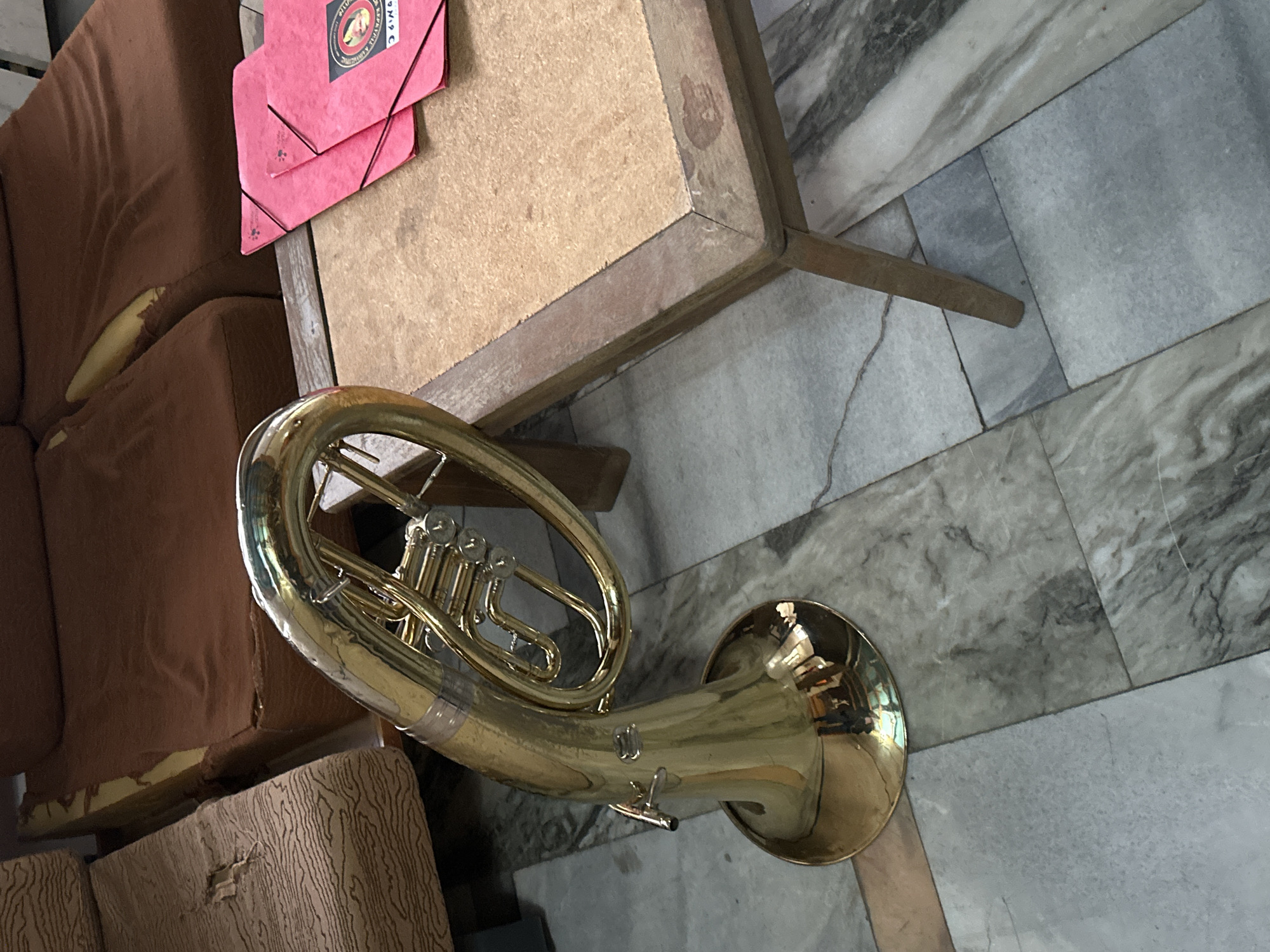 A gold-colored euphonium is positioned on a marble-tiled floor in an indoor setting. The instrument lies near a brown upholstered couch and a wooden table. A red folder sits on the table. The location is unknown.