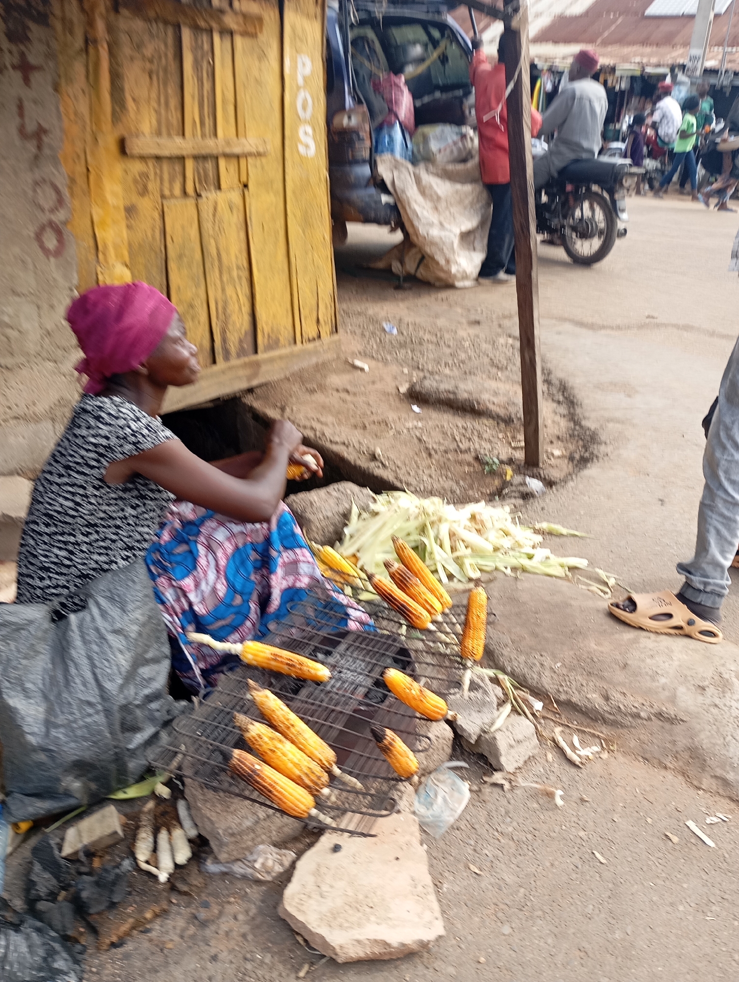 A street vendor in Shendam, Nigeria is grilling corn on a metal rack over coals. The vendor, a woman wearing a pink headscarf and a black and white patterned dress, is seated next to the grill. She is holding an ear of corn in her right hand, and it appears she is in the process of preparing it for grilling. The corn is arranged on the rack, and some pieces are already showing signs of charring. In the background, there is a yellow wooden structure with the letters "POS" written vertically in white. Behind that, a busy market scene unfolds with people and vehicles. The ground is a mix of dirt and pavement, with some debris scattered around. The overall impression is that of a vibrant, bustling market in Nigeria.