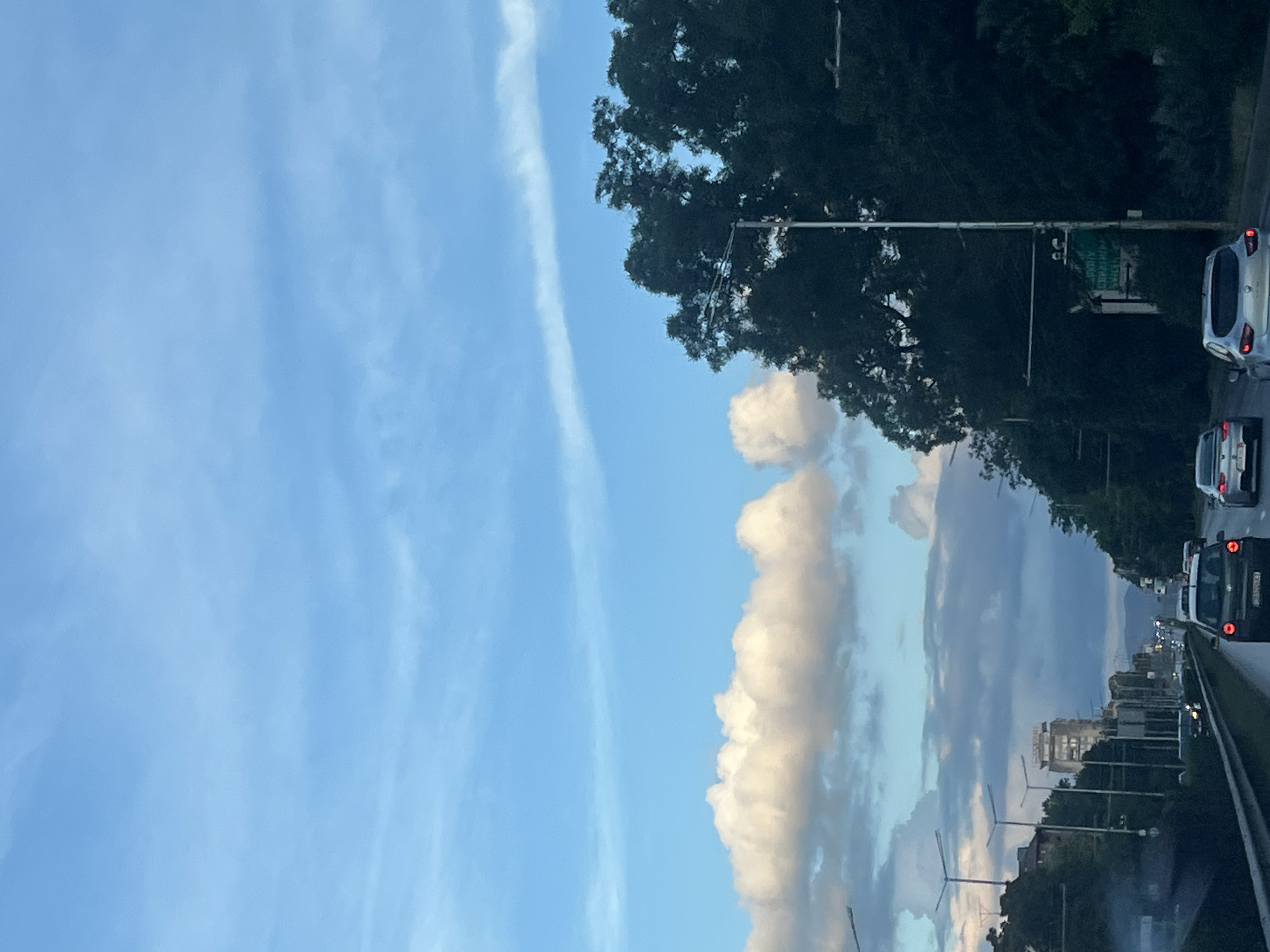 The image, oriented vertically, depicts a road scene with a clear blue sky on the left and a highway on the right. The sky is characterized by scattered white and grey clouds, with a notable, wispy cirrus cloud stretching across the upper left quadrant. Dense green trees line the right side of the highway, with a bright white pole featuring a sign structure visible above the trees. A silver car is seen on the right, with its brake lights illuminated, suggesting traffic is present. Further down the road, more cars are visible, indicating a busy highway. In the distance, a city skyline with buildings can be seen under a dusky sky, with lights from the city visible in the background. The overall impression is that of a late afternoon or early evening on a partly cloudy day, with a journey underway on a highway. No people are directly visible in the image, but the presence of cars implies human activity. The mood is calm, possibly reflective, with the natural elements of the sky and trees contrasting with the man-made environment of the road and city. There is some text on a green sign above the trees, but it is too blurry to be legible.