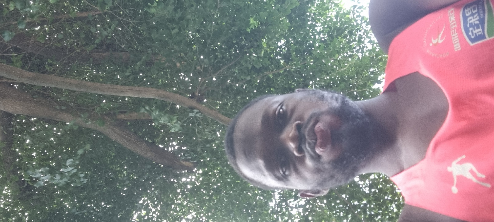 A dark-skinned man with a beard and a pout is taking a selfie outdoors. He is wearing a bright pink tank top with a white graphic of a figure in mid-stride holding a ball on the left side of his chest. The background consists of lush green foliage and tree branches, suggesting a natural, possibly park-like setting within Ikoyi, Nigeria. The lighting is bright, with dappled sunlight filtering through the leaves.
