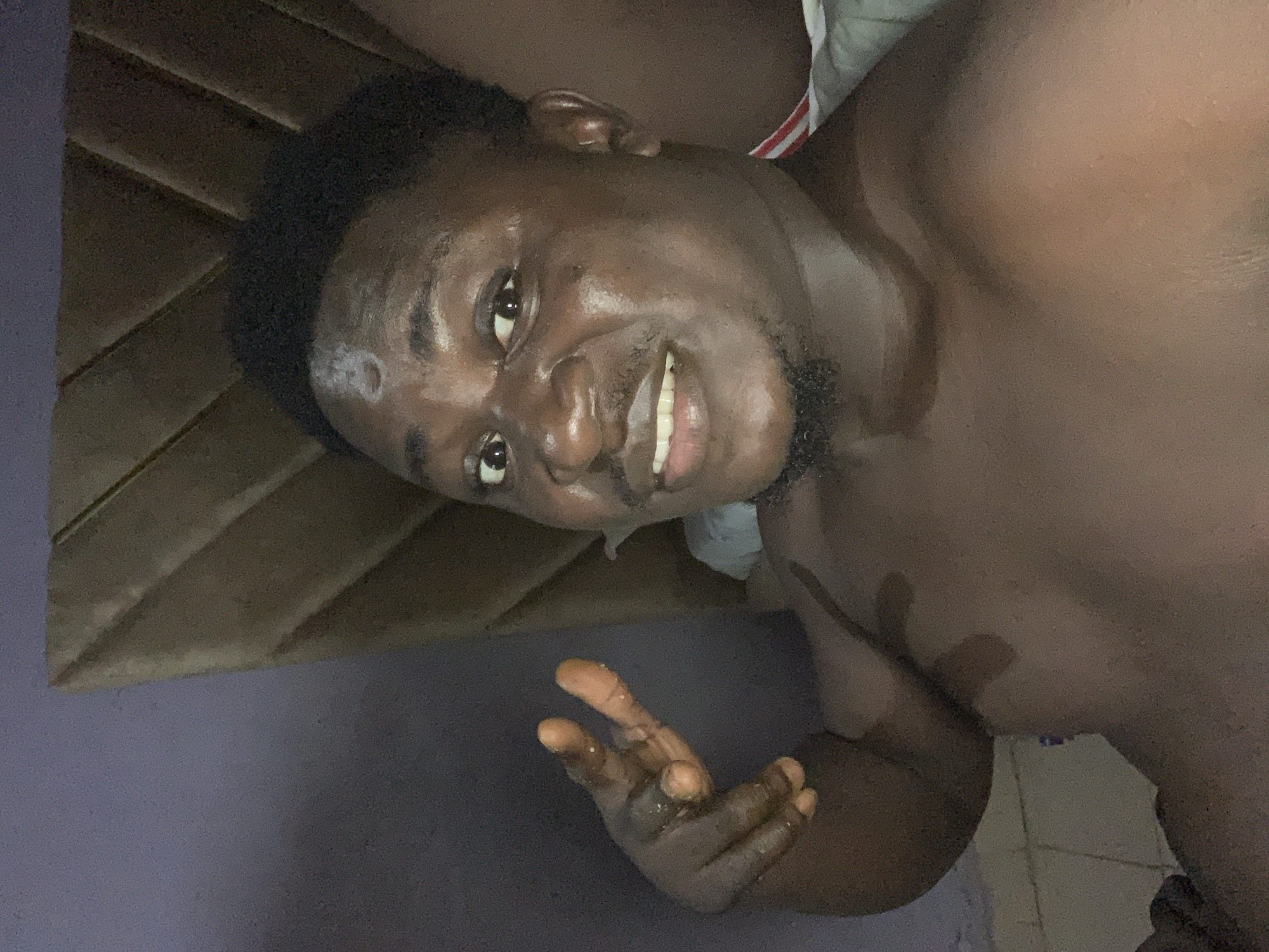 A close-up, indoor, selfie-style photograph shows a Black man with dark skin, dark hair, and a goatee. He is shirtless and appears to be sweating. His right hand is raised, with his index and middle fingers extended in a peace sign. His mouth is open, and he is smiling. The background consists of a textured wall and what appears to be a wooden structure. The lighting is dim, with a bright light source to the left of the frame. The overall impression is casual and personal. The location is specified as Akwanga, Nigeria.
