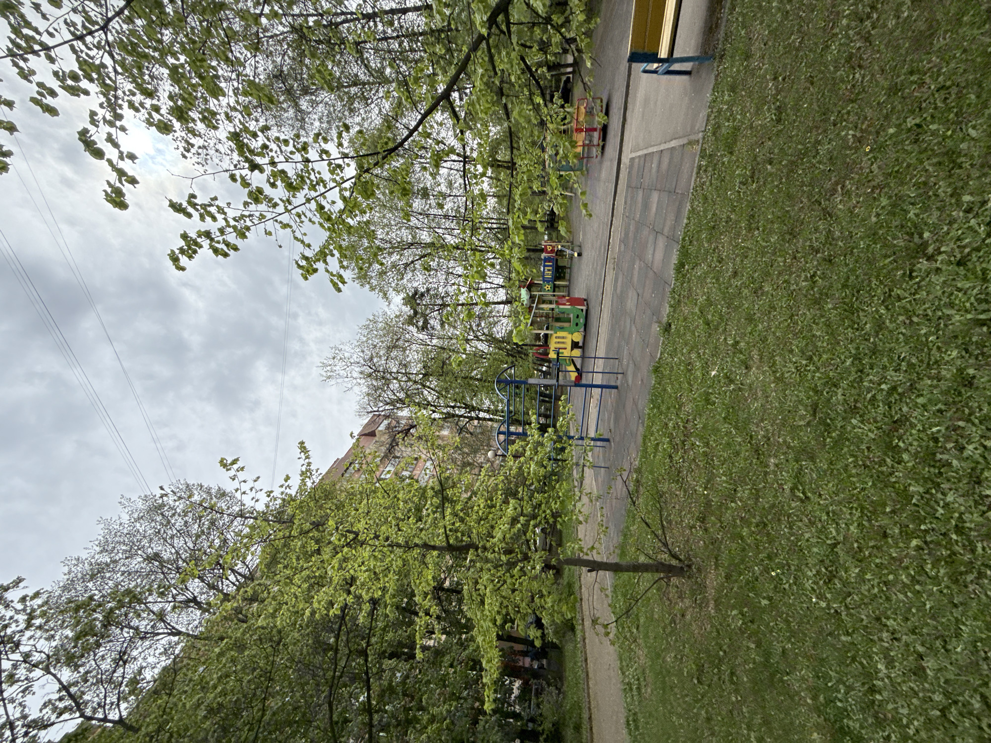 The image shows a playground in Mytishchi, Russia. The playground has several pieces of equipment, including a blue climbing frame and a colorful train. The playground is surrounded by grass and trees with fresh leaves. A sidewalk runs along the side of the playground, with a bench visible. A multi-story building is partially visible in the background, behind the trees. Overhead power lines are visible against a cloudy sky. The overall impression is of a typical urban park setting.