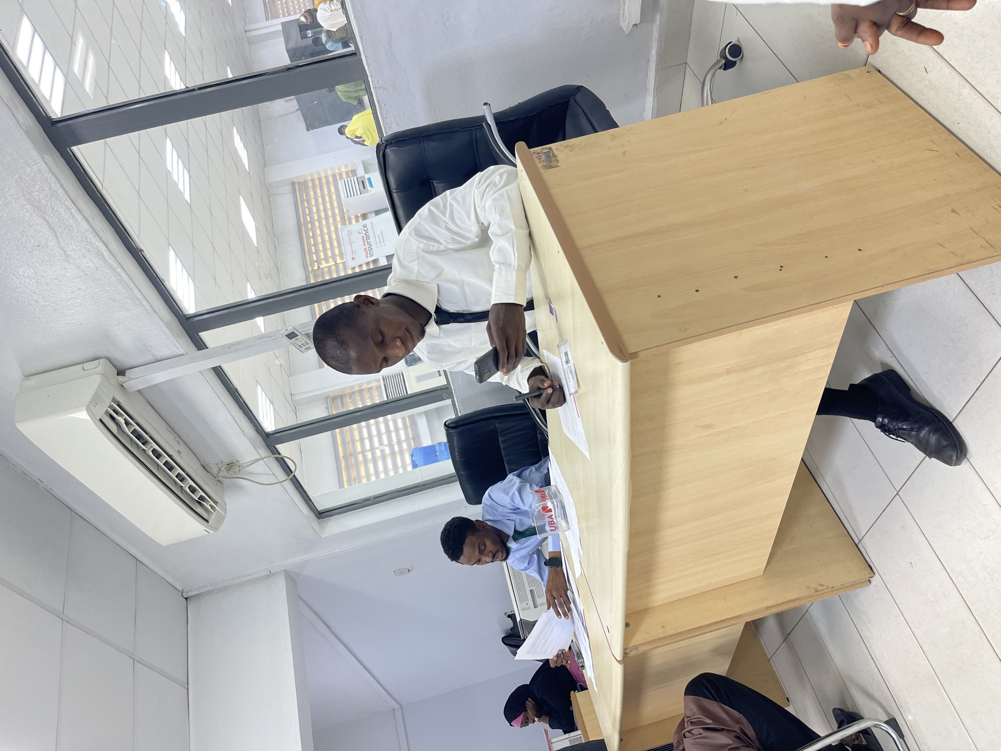 The image captures an indoor scene, likely an office or bank, with several individuals engaged in work-related activities. The perspective is from an elevated, slightly angled position, looking down at two men seated at light wood desks.
In the foreground, a man in a white long-sleeved shirt and dark tie is seated at a desk, looking down at a small object in his left hand (possibly a phone) while his right hand holds a pen, seemingly writing on papers on the desk. He wears dark shoes. Behind him, another man in a light blue collared shirt and dark tie is also seated at a similar desk, looking down at documents. A transparent container with a red and white logo and the letters "UBA" is visible on his desk. To the left and slightly behind this man, a person wearing a dark hijab with a pink trim is partially visible, also appearing to be seated at a desk.
The setting includes large glass windows in the upper part of the wall, allowing natural light into the space and revealing glimpses of other individuals and what appears to be another room or hallway outside. An air conditioning unit is mounted on the wall above the desk where the man in white is seated. The floor is tiled.
Visible text includes the word "Insurance" on a sign seen through the window, and "UBA" on the container on the second man's desk. The overall atmosphere suggests a typical workday in an administrative environment, consistent with a bank or office setting in Zaria, Nigeria.