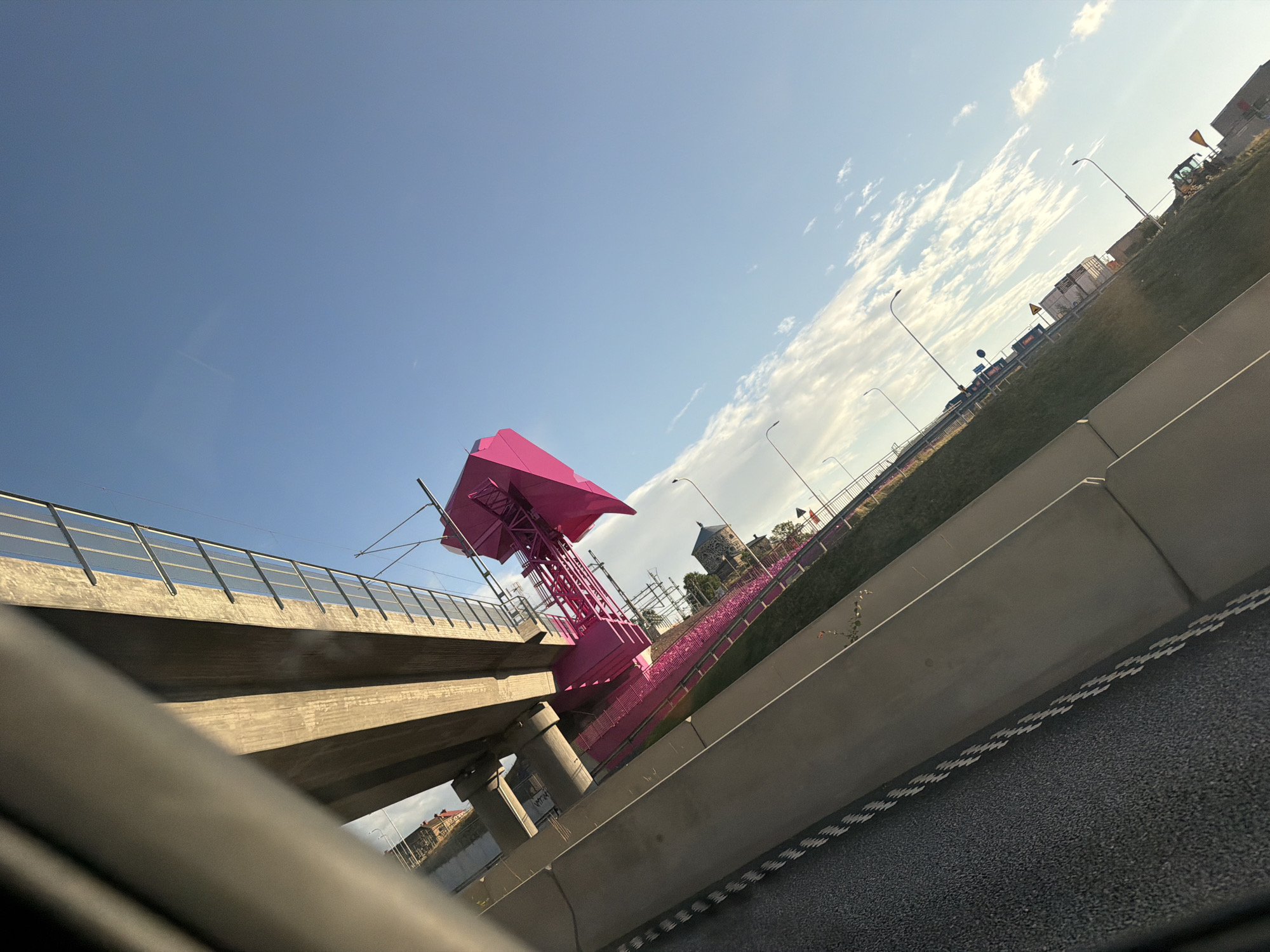 The image, taken from a low angle within a vehicle, shows a brightly colored pink structure, possibly part of a temporary installation or artwork, adjacent to a concrete bridge or overpass. The structure is situated on a grassy embankment alongside a road. The sky is a clear blue with scattered white clouds, and the scene appears to be illuminated by natural daylight. In the background, there are buildings, streetlights, and some construction equipment, including a yellow excavator. The overall impression is of an urban or semi-urban environment with recent construction or public art. This structure is likely located in Gothenburg, Sweden, as it is a well-known pink art installation that was part of the city's harbor revitalization.