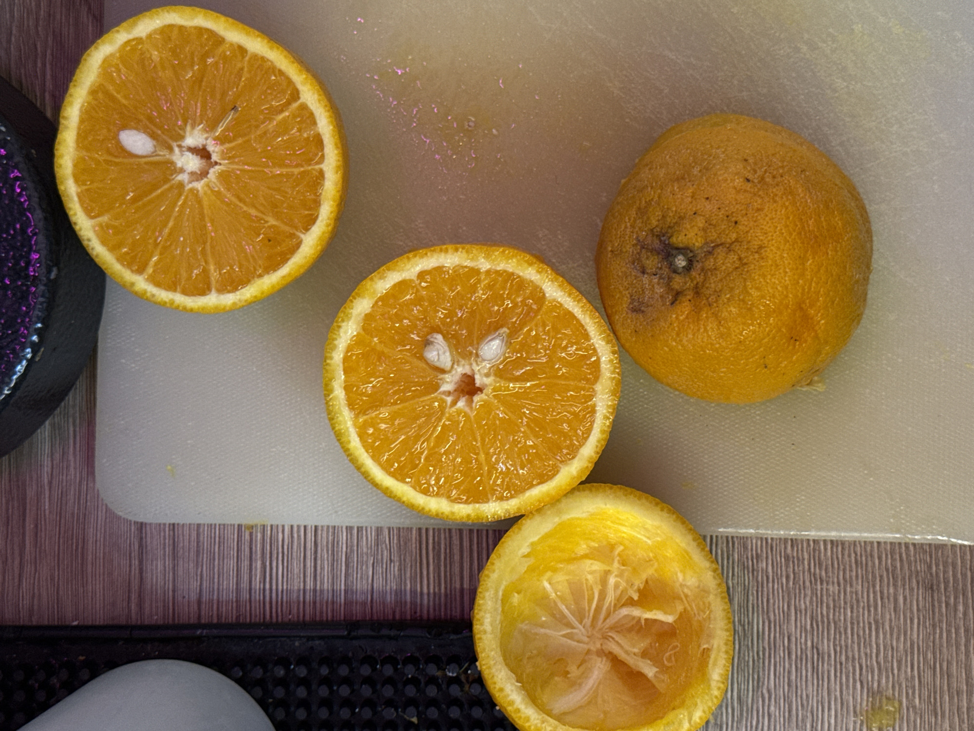 The image shows four oranges on a white cutting board and a wooden surface. One whole orange is positioned in the lower right quadrant, with visible blemishes on its skin. To its left, on the cutting board, is a halved orange, its interior facing upwards, revealing segments and seeds. Above this halved orange, also on the cutting board, is another halved orange, oriented differently, showcasing its juicy interior and a prominent seed. To the left of the cutting board, on a wooden surface, is the squeezed peel of an orange. The lighting suggests an indoor setting. The image does not provide any information or visual cues that would allow for its location to be identified as Moscow, Russia.