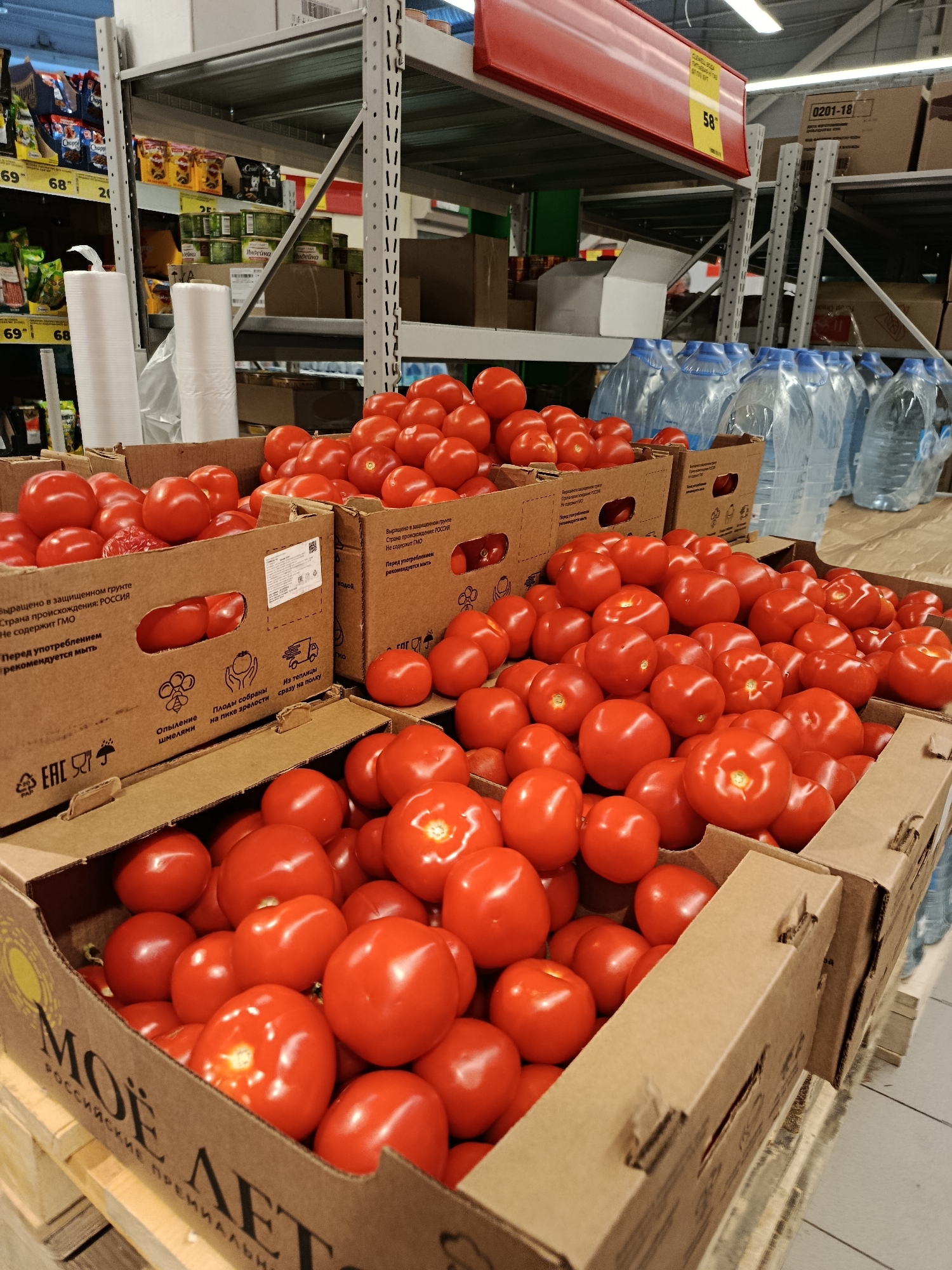 This is a medium shot of a produce section in a supermarket, likely in Orekhovo-Zuyevo, Russia. The main subjects are boxes and crates filled with ripe red tomatoes. The tomatoes are the primary focus, displayed in bulk on shelves and in cardboard display boxes. In the background, shelves are stocked with various food items, including canned goods and bottled water.
The scene appears to be set during daytime, as indicated by the bright artificial lighting common in retail environments. There are no people visible in the immediate foreground, suggesting the photograph was taken during a quiet period or when staff were restocking. The overall mood is neutral and commercial, highlighting the freshness and abundance of the produce.
Notable details include the prominent display of multiple boxes of tomatoes, some with labels in Russian. One visible box has text that translates to "Grown in a protected ground. Country of origin: Russia. Does not contain GMO. Recommended to wash before consumption." Another box features branding that reads "MOЁ ЛET" and "RUSSIAN PREMIUM". Signs on the shelves indicate prices, such as "69" and "68", and a yellow price tag with "58" on it. The setting is clearly a supermarket aisle, with metal shelving units and cardboard pallets visible. The tomatoes themselves are round and vibrant red, suggesting they are of good quality. The arrangement of the tomatoes and the packaging imply a retail setting where fresh produce is being sold.
