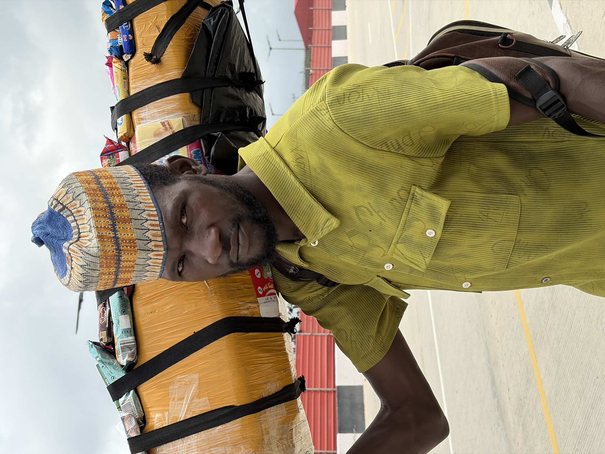 A dark-skinned man wearing a green corduroy shirt and a colorful embroidered cap carries a large, yellow-wrapped package on his shoulder. The package is secured with black straps and has various small, individually wrapped items on top, including what appear to be candy bars or snacks. He has a beard and is looking directly at the camera. A brown backpack is slung over his left shoulder. The background suggests an outdoor urban environment, possibly an airport or a large open area with paved ground marked with yellow lines. In the distance, there are red and white striped barriers and light poles, indicating infrastructure typical of a transport hub or a modern city area. The sky is overcast. The image was taken in Igboefon, Nigeria.