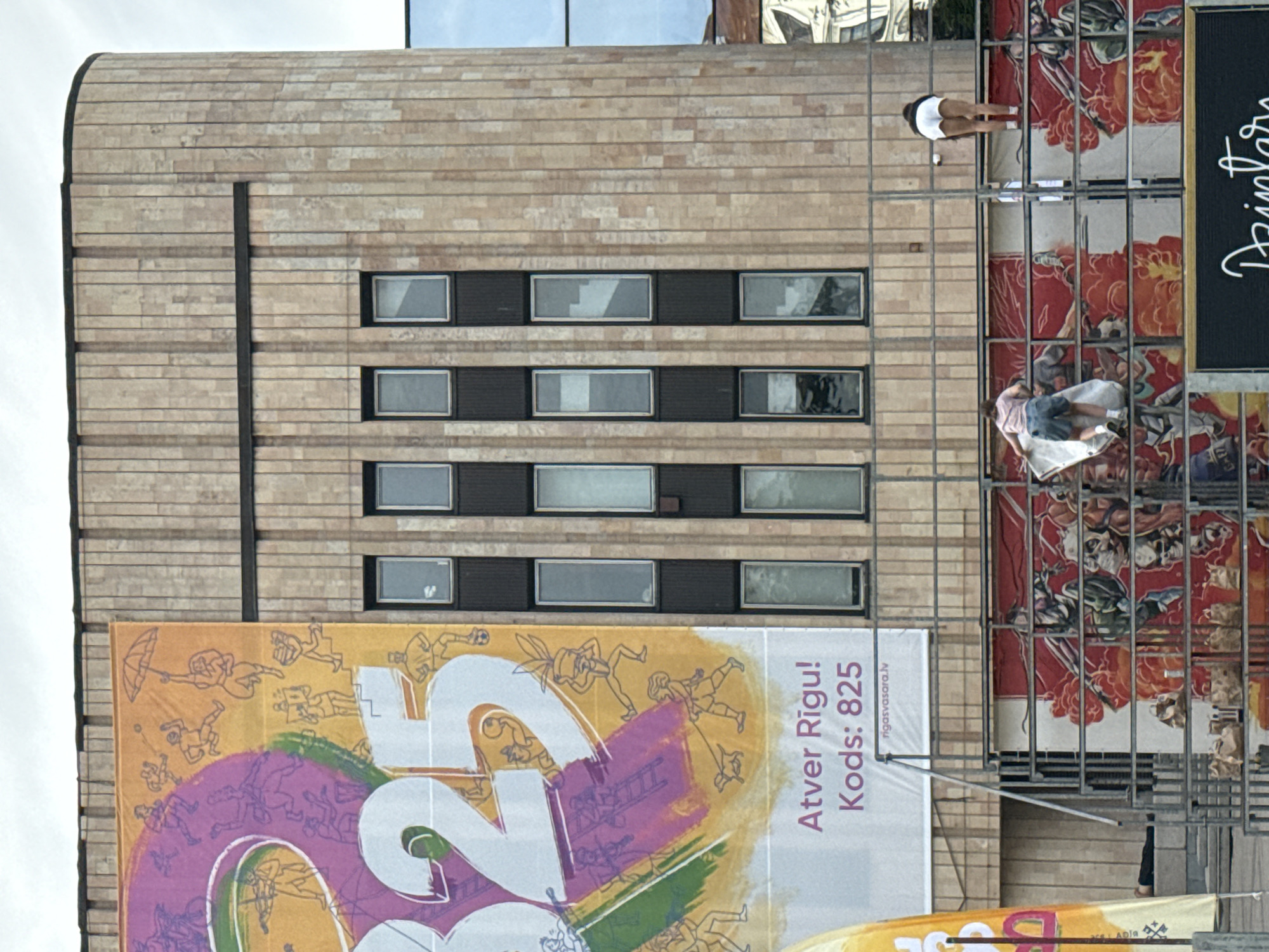 The image depicts a street scene in Riga, Latvia, focusing on a large building facade adorned with a colorful banner and a mural. The building has a beige, vertically paneled stone exterior with several dark-framed windows arranged in vertical rows. A large, vibrant banner, partially visible on the left side of the building, features large white numerals "325" and an illustration of various stylized figures engaged in activities like playing music, sports, and holding an umbrella. Below the banner, text reads "Atver Rigu! Kod's: 825" and a website address "rigasvasara.lv".
In the foreground, a metal scaffolding structure runs horizontally across the scene. Behind the scaffolding, a mural with a predominantly red and white color scheme is visible, depicting dynamic figures, some appearing to be athletes or warriors. Several people are interacting with this mural and the scaffolding. On the left, a person in a pink t-shirt and denim shorts is leaning over the scaffolding, holding a white sheet or banner. To the right, a person wearing a white t-shirt and white shorts is bending over, facing away from the camera, with their back to the viewer. Their posture suggests they are either adjusting something on the mural or scaffolding, or are in the process of climbing. The overall environment appears to be an urban setting, likely a public space given the banner and mural.