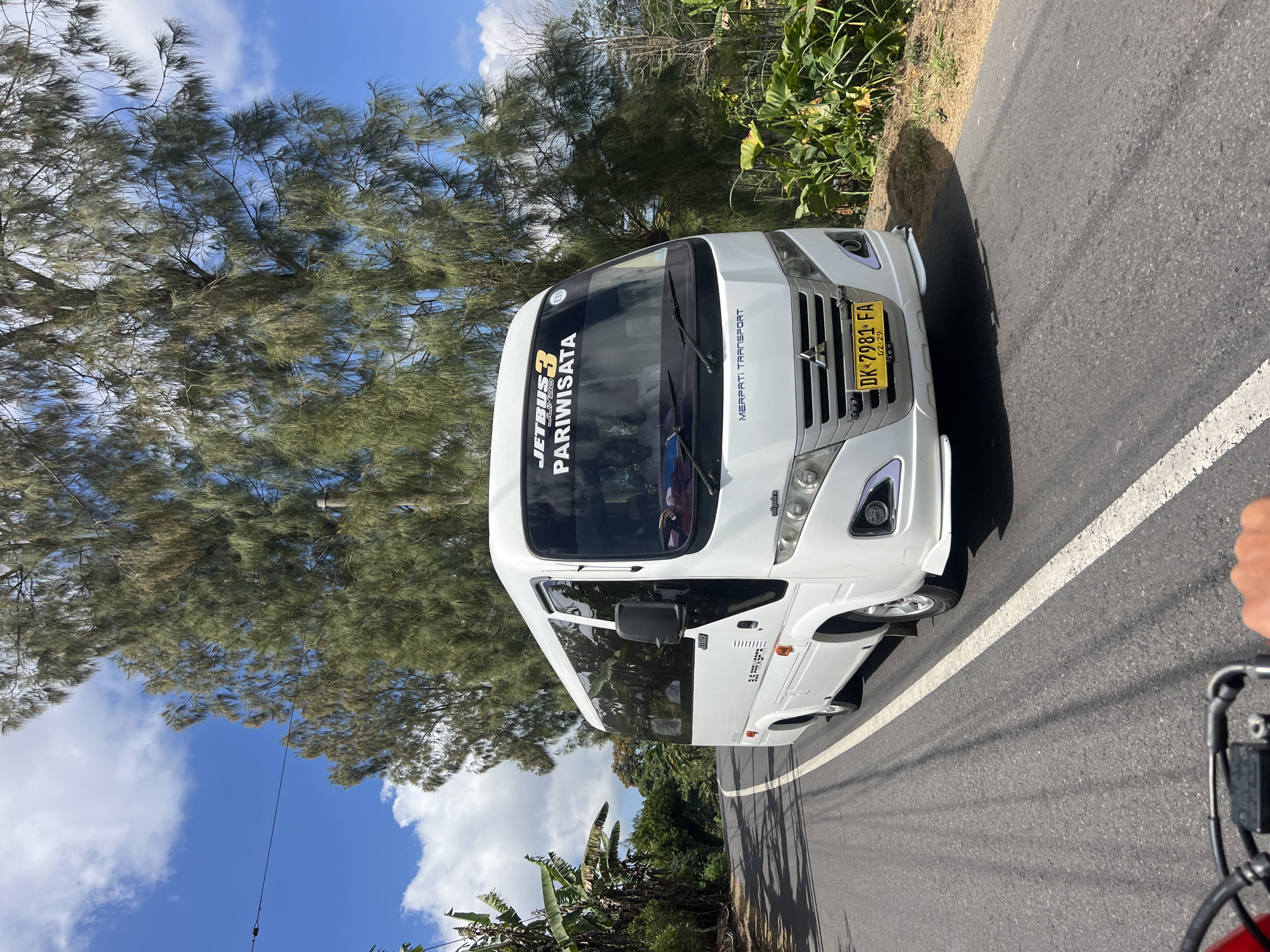 A white passenger bus with "JETBUS 3 PARIWISATA" printed on its windshield is driving on a paved road in Kintamani, Indonesia. The bus has a license plate that reads "DK 7981 FA". The road is bordered by lush green vegetation and tall trees with needle-like leaves. The sky is blue with scattered white clouds. The scene is captured from the perspective of someone riding behind the bus, possibly on a motorcycle, as indicated by the handlebars visible in the foreground. The bus appears to be in motion, traveling along a winding road.
