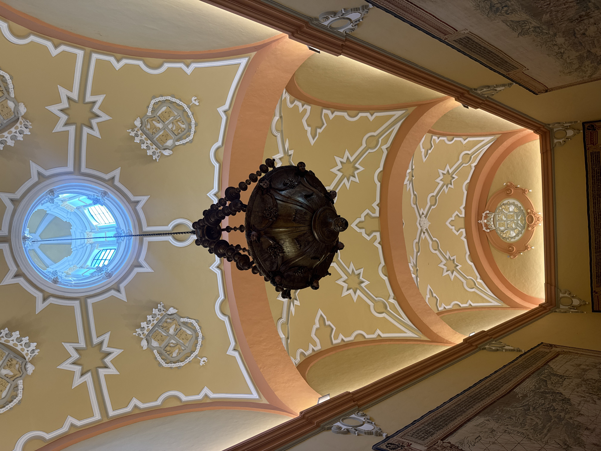 This is a low-angle, interior shot of a hallway ceiling in the Alcázar of Seville, Spain. The ceiling is ornately decorated with a yellow background, accented by white stucco detailing and orange curved beams. A large, dark, antique chandelier hangs from the center, suspended by a chain. In the distance, a circular stained-glass skylight is visible. Along the walls, large tapestries are displayed. The overall impression is one of grandeur and historical richness.