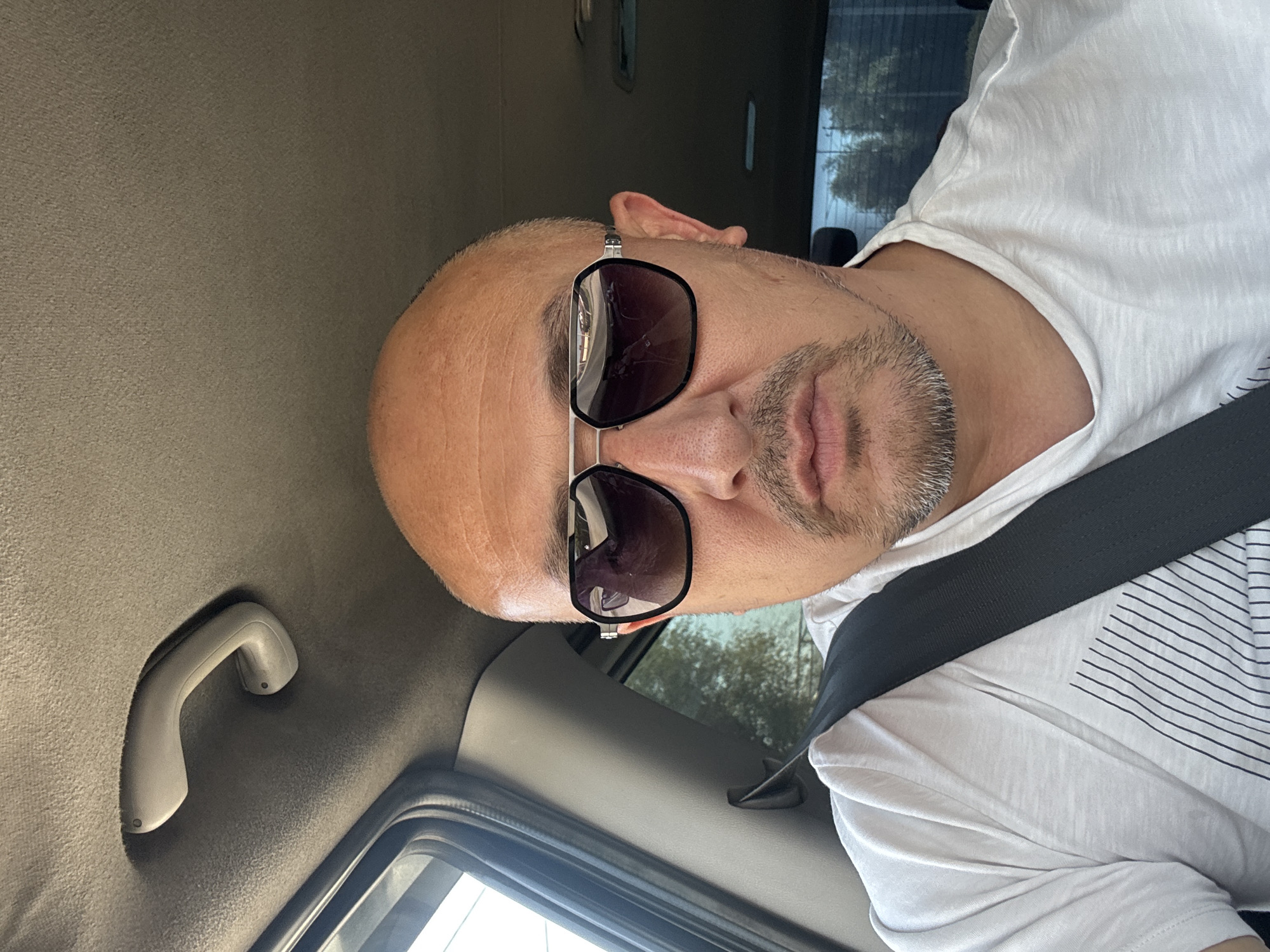 The image shows a man wearing sunglasses and a white t-shirt, seated in the driver's seat of a car. He has a bald head and a goatee. The car's interior is visible, including a handle on the ceiling and a seatbelt. The background outside the car appears to be a street scene with trees and buildings, suggesting an urban environment. The image was taken in the city of Shymkent, Kazakhstan.
