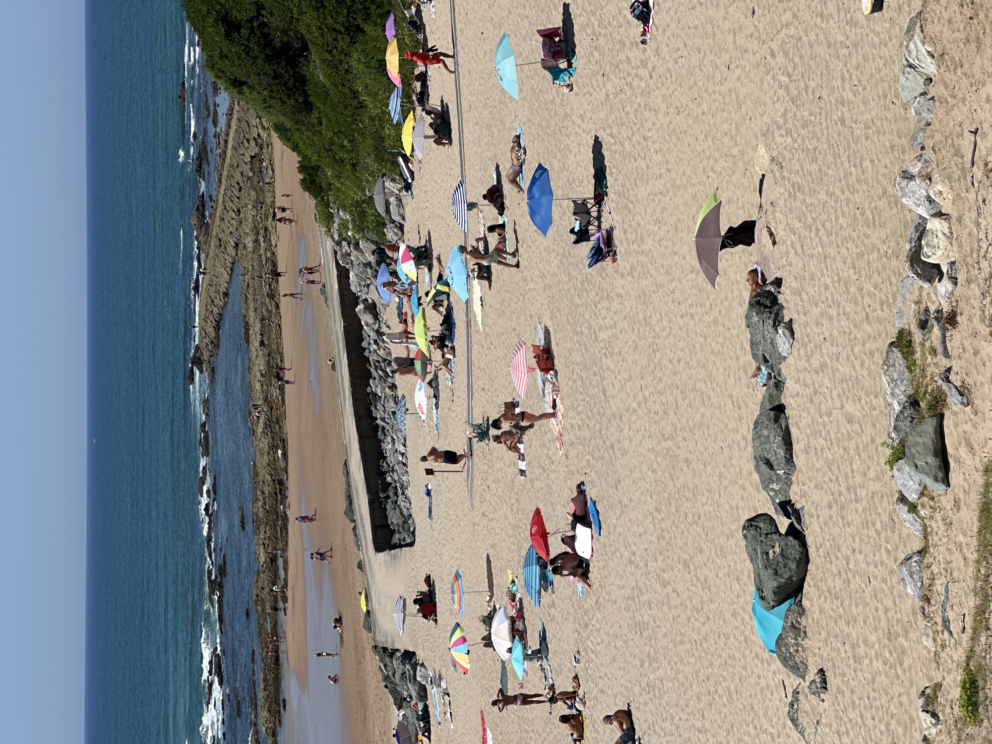 A high-angle, full shot captures a crowded beach scene in Biarritz, France, on a sunny day. The sandy expanse is dotted with numerous beachgoers relaxing under a variety of colorful umbrellas, some of which are large and spread out, while others are smaller and more portable. People are seen sunbathing on towels, reading, and interacting with each other. Some individuals are wading in the shallow water near the shore, and a few are playing in the sand. The ocean, a vibrant blue, stretches out to the horizon, with a rocky breakwater extending into the sea. A lush green hillside borders the beach on the right side of the frame. The overall impression is one of a bustling summer day at the seaside.