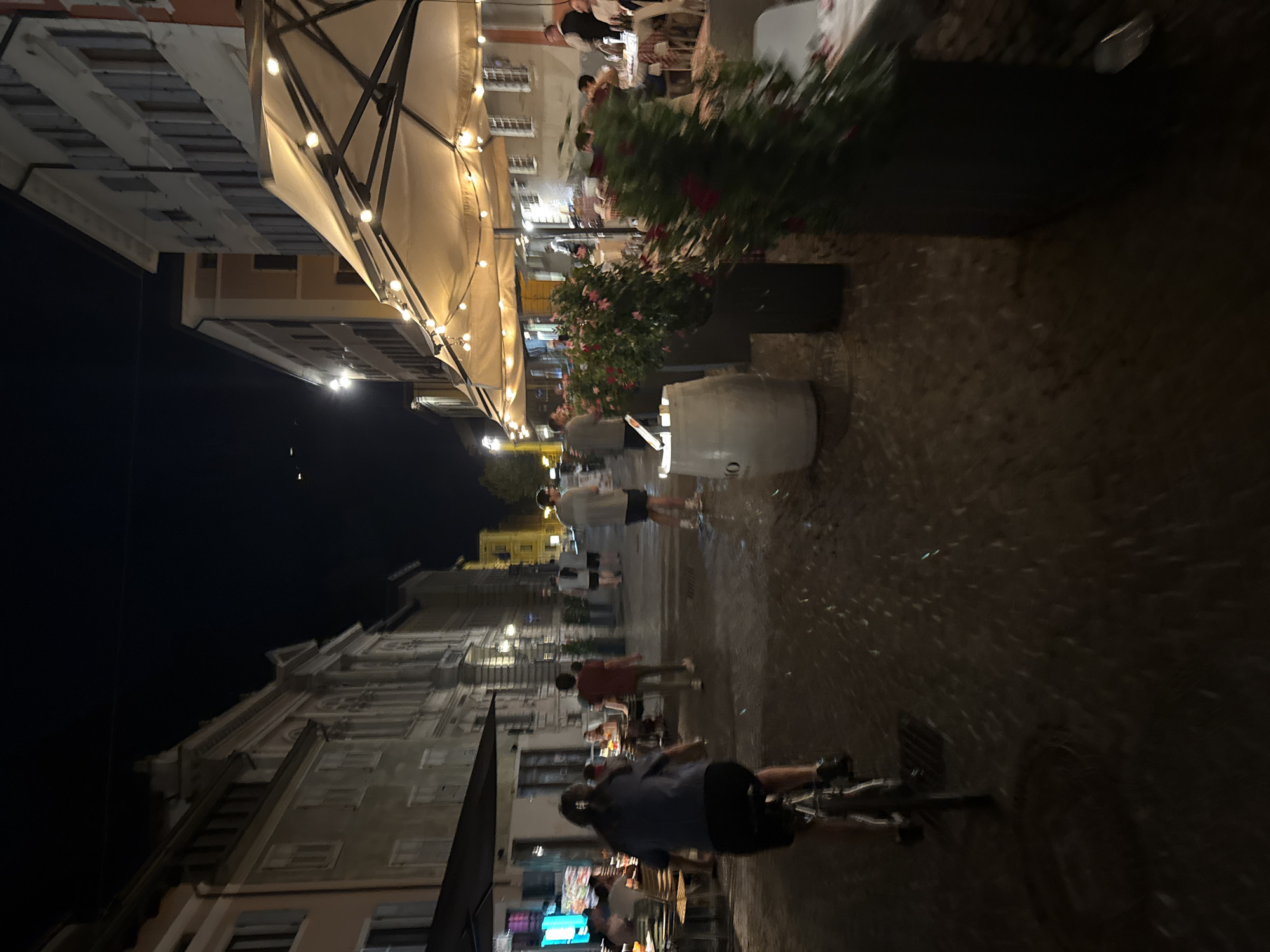 The image depicts a nighttime street scene in Trento, Italy, likely in the historic center. A person is cycling away from the camera on a cobblestone street, illuminated by streetlights and the warm glow of outdoor dining lights. To the right, a restaurant with large umbrellas and strings of lights is set up, with patrons seated at tables. To the left, more buildings line the street, some with their windows lit. The overall atmosphere is that of a lively evening in a European city.