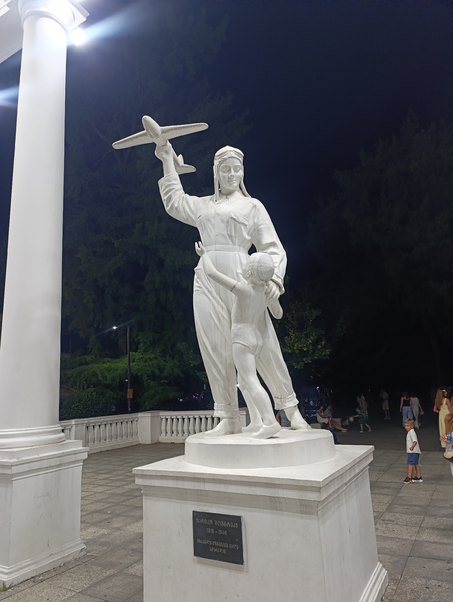 The image shows a white statue of a woman pilot holding a toy airplane and a child. The statue is located in Batumi, Georgia, in front of a white column and a balustrade. The statue stands on a pedestal with an inscription in Georgian. In the background, there are trees and people walking. The scene is illuminated by artificial lights, suggesting it is nighttime. The statue appears to be a monument to a historical figure.