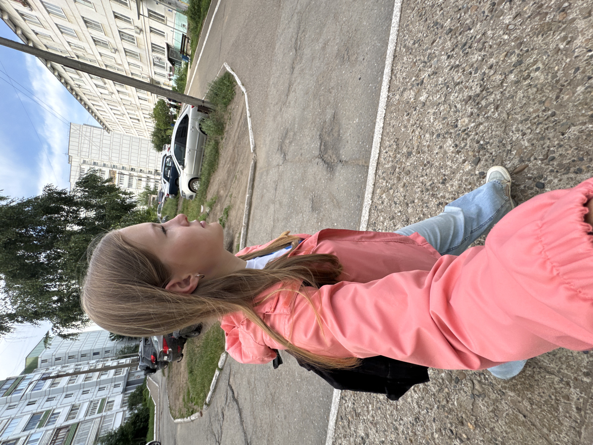 The image shows a young Caucasian female walking on a paved sidewalk in an urban environment. She has blonde hair and is wearing a pink jacket, a white shirt, and light blue jeans. A black backpack is visible on her back. The background features multi-story apartment buildings, trees, and parked cars, consistent with a residential area in Chaikovsky, Russia. The pavement is cracked and uneven, and the weather appears overcast. The perspective suggests the photo was taken by the subject herself, likely with a smartphone.