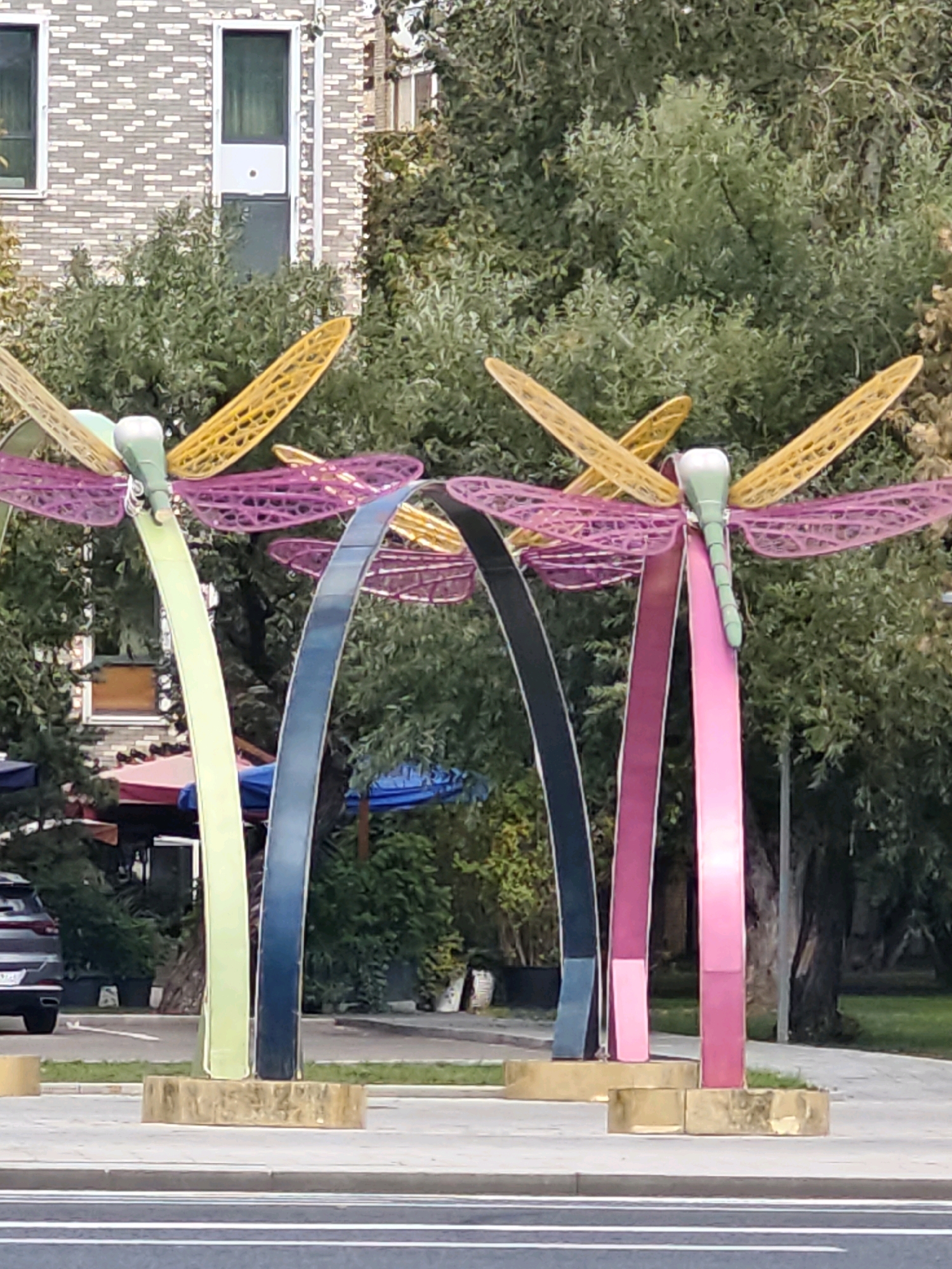 The image depicts an outdoor public art installation featuring four stylized dragonfly sculptures. The dragonflies have large, intricate wings in yellow and pink, and their bodies are a light green. Each dragonfly is supported by a slender, arching structure, with the supports colored in light green, dark blue, and two shades of pink. These sculptures are placed on a paved area, likely a sidewalk or plaza, with a street visible in the foreground. In the background, there is a brick building, lush green trees, and some street furniture, including awnings and planters. A grey car is parked to the left of the sculptures. The scene appears to be in an urban environment. This artwork is located at the "Dragonfly" residential complex on Marshala Zakharova Street in Moscow, Russia.