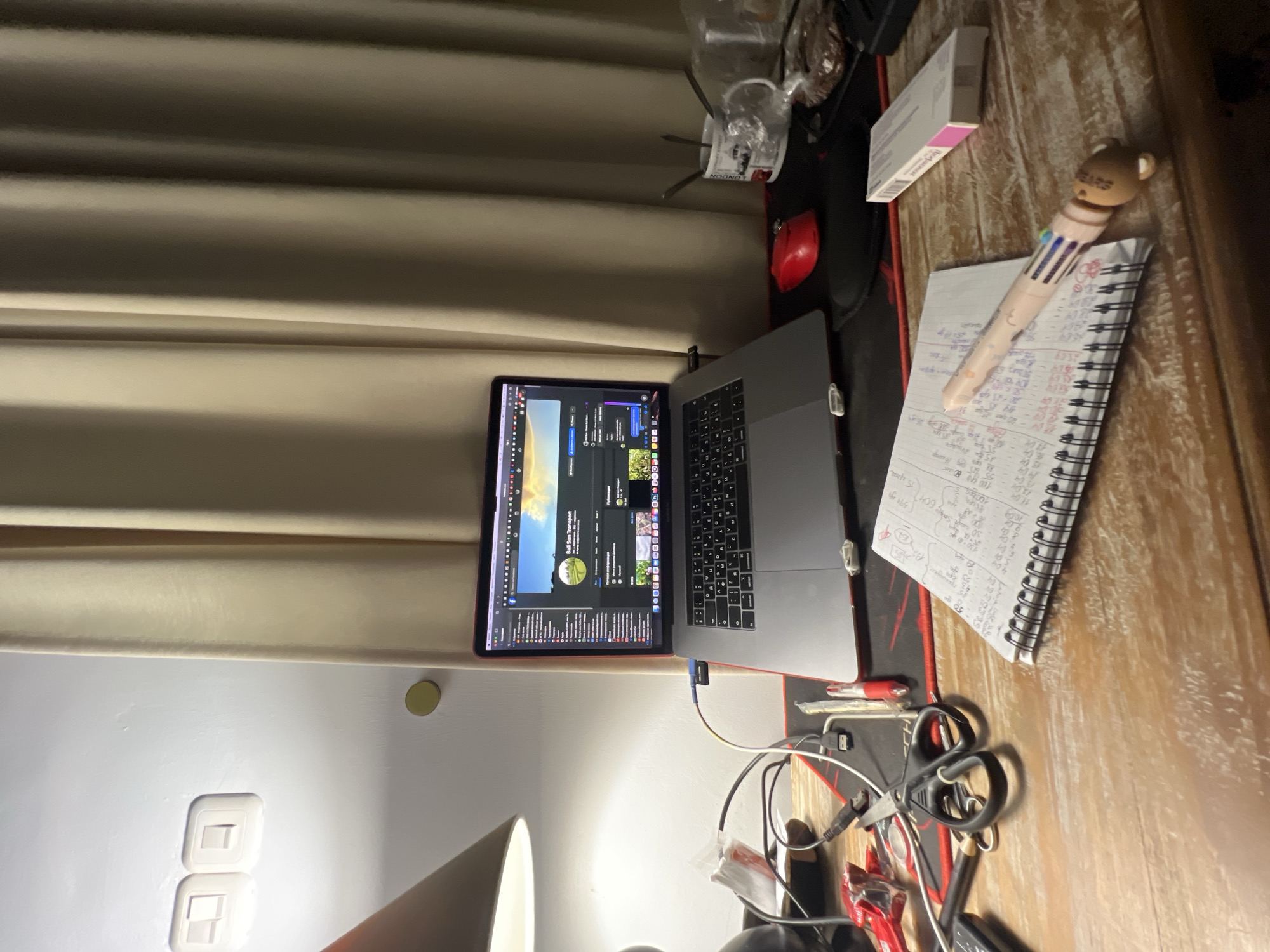 An overhead, slightly angled shot captures a desk setup in what appears to be an indoor setting, possibly an office or study area, in North Kuta, Indonesia. The dominant items on the wooden desk are an open MacBook Pro laptop and a spiral-bound notebook.
The laptop's screen displays a web page, likely a social media platform or business profile, with "Bali Sun Transport" clearly visible at the top, along with various icons and user interface elements. Next to the laptop, a black and red mousepad is partially visible, with cables and a pair of black-handled scissors lying on it.
To the right of the laptop, a white spiral notebook is filled with extensive handwritten notes, primarily numbers and calculations, some of which are crossed out or underlined. Visible text includes "6354", "7229399", "10.09", "11.04", "12.04", "13.04", "50", "130", "5944", "30 S 604 15", "1000", "100 460", "55 256", "88", and "15". Resting on the notebook is a multi-color pen adorned with a small bear head topper.
Further back on the desk, near the curtains, there's a small white box, an empty glass or cup with the word "LONDON" printed on it, and a small red object, possibly a computer mouse or a small device. Beige or cream-colored curtains are visible in the background, suggesting a window or partition. To the lower left, part of a white wall with two light switches or outlets is illuminated by a desk lamp, whose shade is partially visible at the bottom of the frame. The overall impression is an active, somewhat cluttered workspace, likely used for business or administrative tasks.