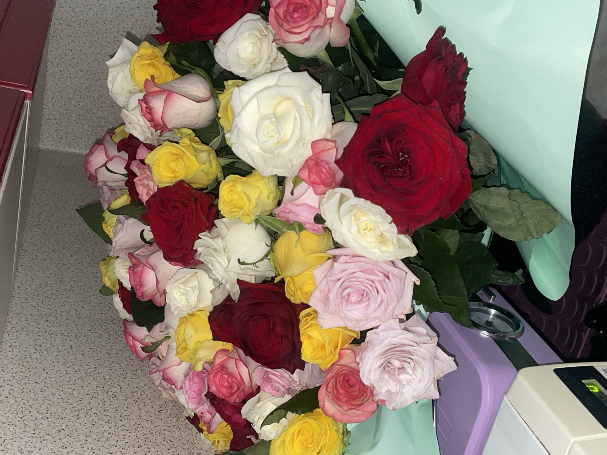 This close-up image features a vibrant, multi-colored bouquet of roses, viewed from a slightly elevated angle. The bouquet is densely packed with a variety of rose colors including deep red, bright white, sunny yellow, and various shades of pink and light purple. Some roses exhibit bi-colored petals, with subtle transitions from one hue to another. Green leaves are visible among the blossoms, providing natural contrast.
The bouquet is partially wrapped in a soft mint green or pale blue material, which is most prominent on the right side. The background indicates an indoor setting. To the left, a reddish-brown object with a silver accent is visible. The surface supporting the bouquet is a light grey material with dark speckles, possibly a countertop or textured floor. On the lower right, a purple object with a silver ring and a beige electronic device with a small green light are partially visible. The lighting appears to be artificial and somewhat soft, creating gentle shadows. No specific human activity is depicted, but the bouquet suggests a gift or display. There is no visible text in the image.