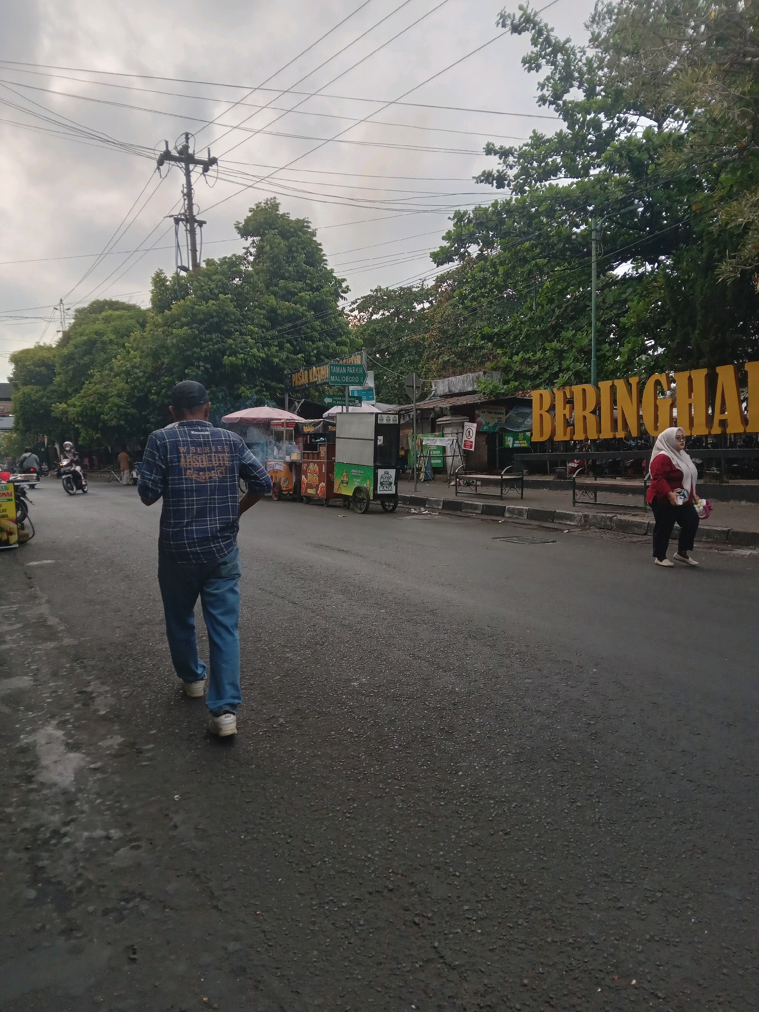 This is a medium shot of a street scene in Yogyakarta, Indonesia, likely in the area of Malioboro or near a park. A man wearing a blue checkered shirt and jeans walks away from the camera on the left side of the road. He is wearing a black baseball cap. On the right side of the road, a woman wearing a red hijab and maroon top with dark pants walks towards the camera. Behind her, a large yellow sign with the word "BERINGHARJO" is visible, indicating a market or a well-known landmark. Several food carts are lined up on the right side of the street, suggesting a commercial or food stall area. Trees line the background, and power lines crisscross the overcast sky. The road surface is asphalt, with some damp patches. Vehicles, including motorcycles, are present on the road. The overall impression is a busy, casual street in a city, possibly during the late afternoon or early evening due to the lighting conditions.