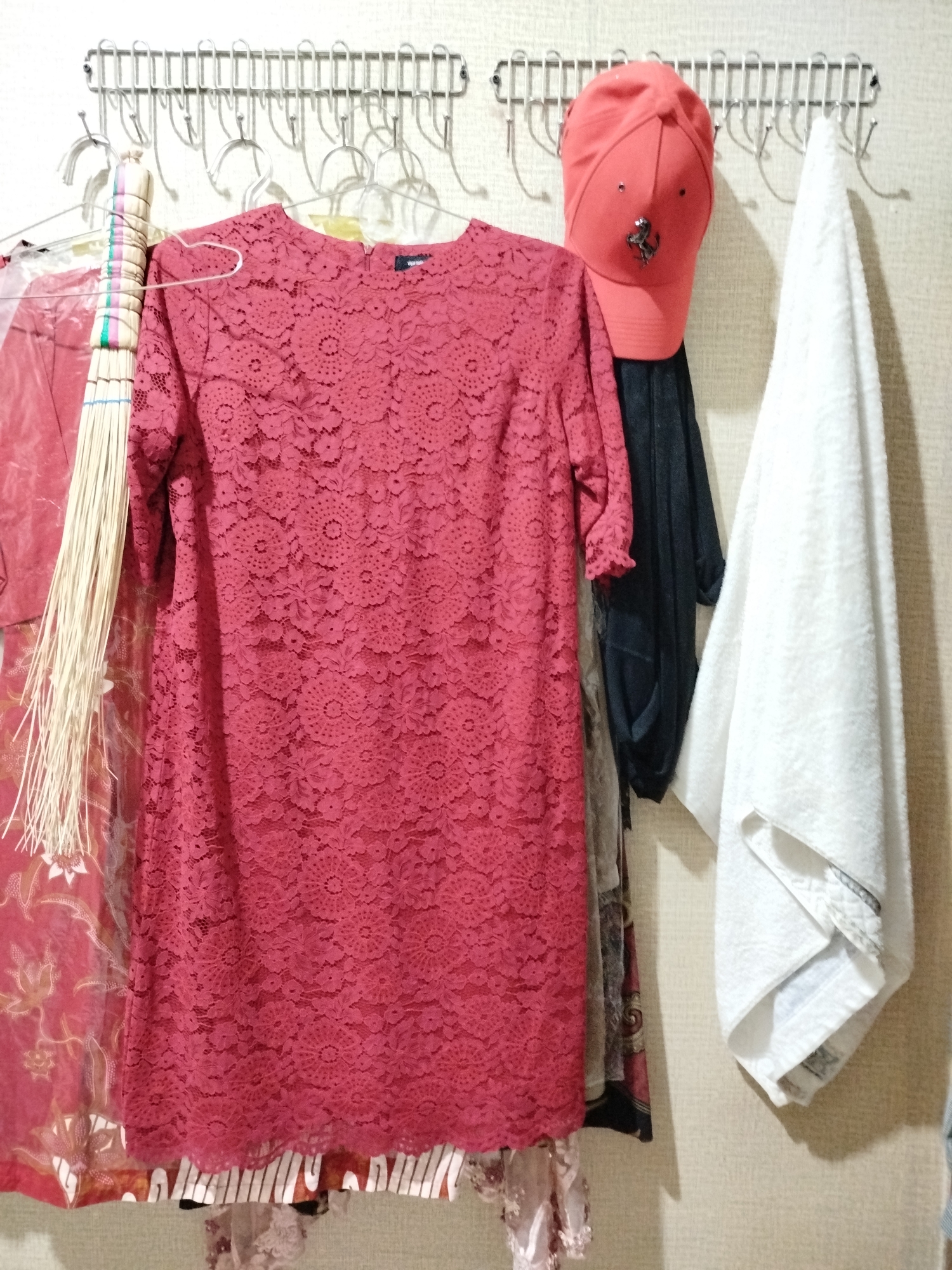 The image depicts a wall with a metal rack holding various items. On the rack, there are at least two garments: a red lace dress and a dark-colored garment, possibly a shirt or jacket, hanging behind it. To the left of the red dress, there is a broom with a light-colored handle and bristles. A red baseball cap with a metallic emblem is hanging to the right of the dark garment. Further to the right, a white towel is draped over a hook. The background appears to be a textured wall. The image does not provide enough information to determine the specific location within Bandungrejo, Indonesia.