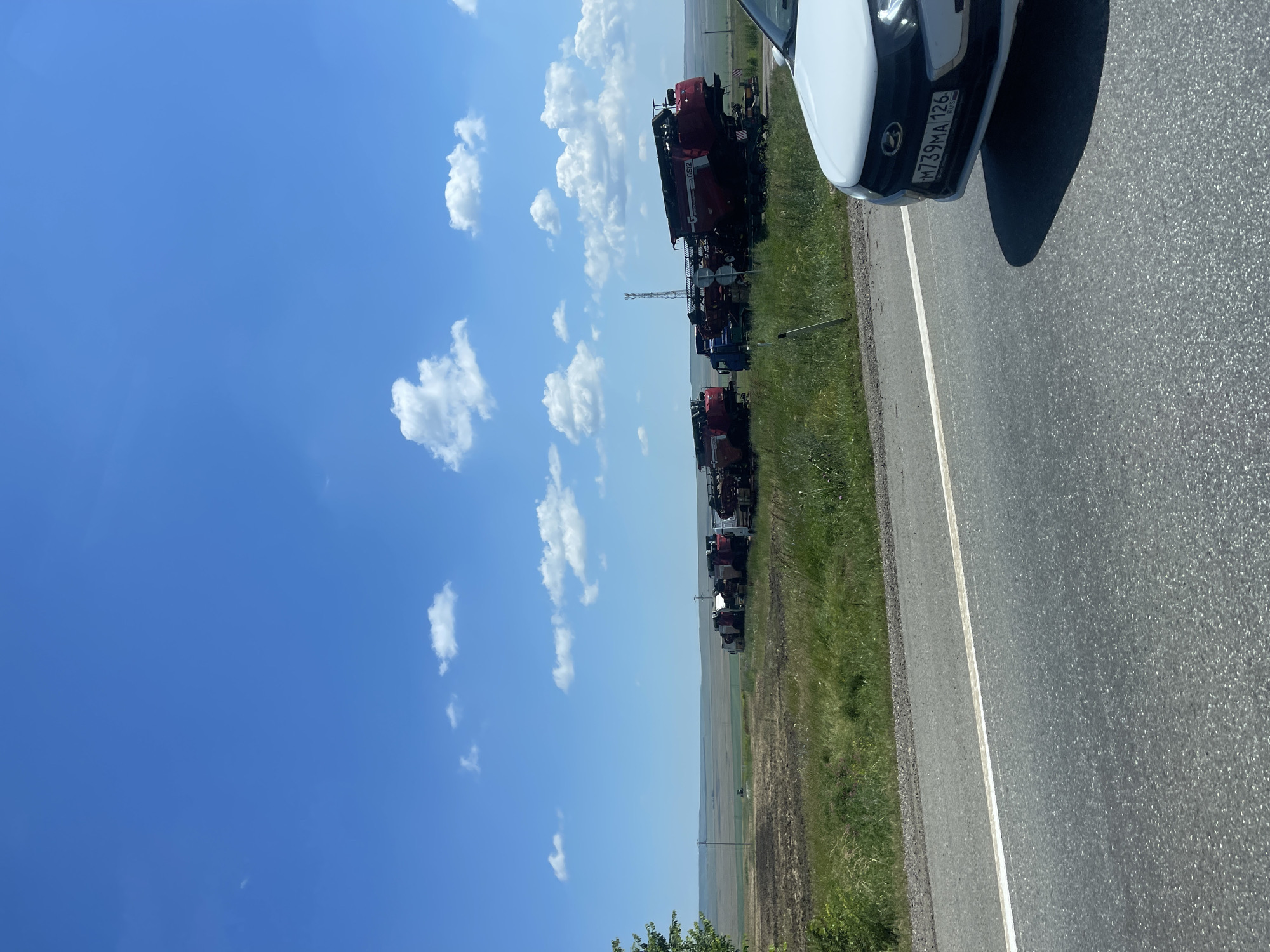 The image shows a roadside scene in Kursavka, Russia. The foreground is dominated by the right side of a white car, revealing its front end and license plate. The license plate reads "M739MA 126". Adjacent to the car is an asphalt road with a white lane marking. To the left of the road, a grassy field is visible, with some agricultural fields in the distance. Several large red agricultural machines, likely harvesters or combines, are parked in a line along the side of the road. These machines are the main focus of the composition, stretching from the mid-ground into the background. Above the landscape, a bright blue sky with scattered white cumulus clouds fills the upper portion of the image. The sunlight suggests it is daytime, likely late morning or early afternoon, given the clear skies and shadows. The scene appears calm and rural, with no people visible. The overall impression is one of agricultural machinery staged for transport or awaiting use in the fields.