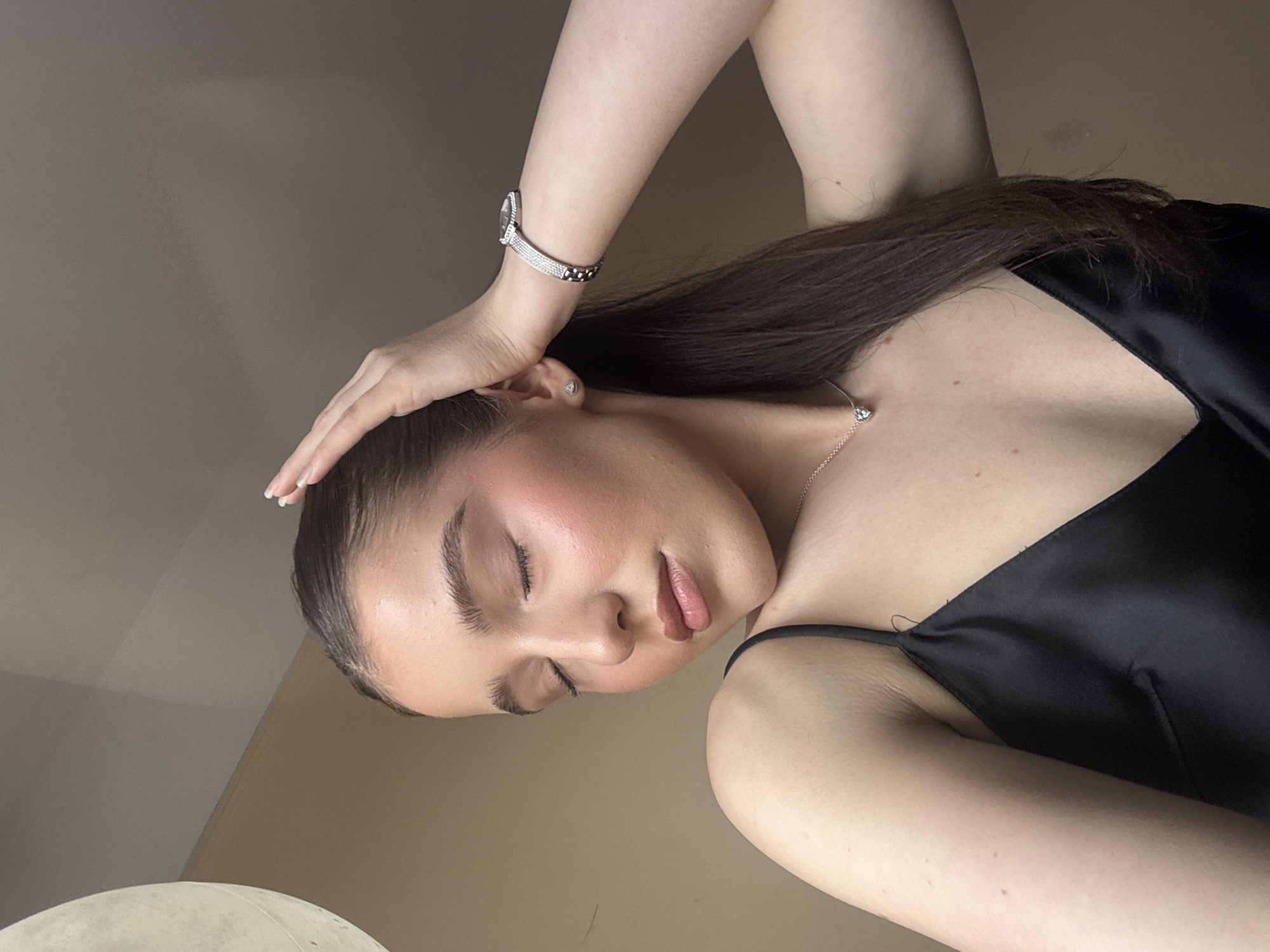 A woman is positioned at an oblique angle with her head resting on her hand. She is wearing a black silk camisole, and a delicate silver necklace is visible around her neck. A silver watch is on her wrist. Her eyes are closed, and her facial makeup is noticeable, including blush on her cheeks and lipstick. The background is a neutral beige wall with some lighting in the lower left corner, possibly from a lamp or window.
The location is unknown.