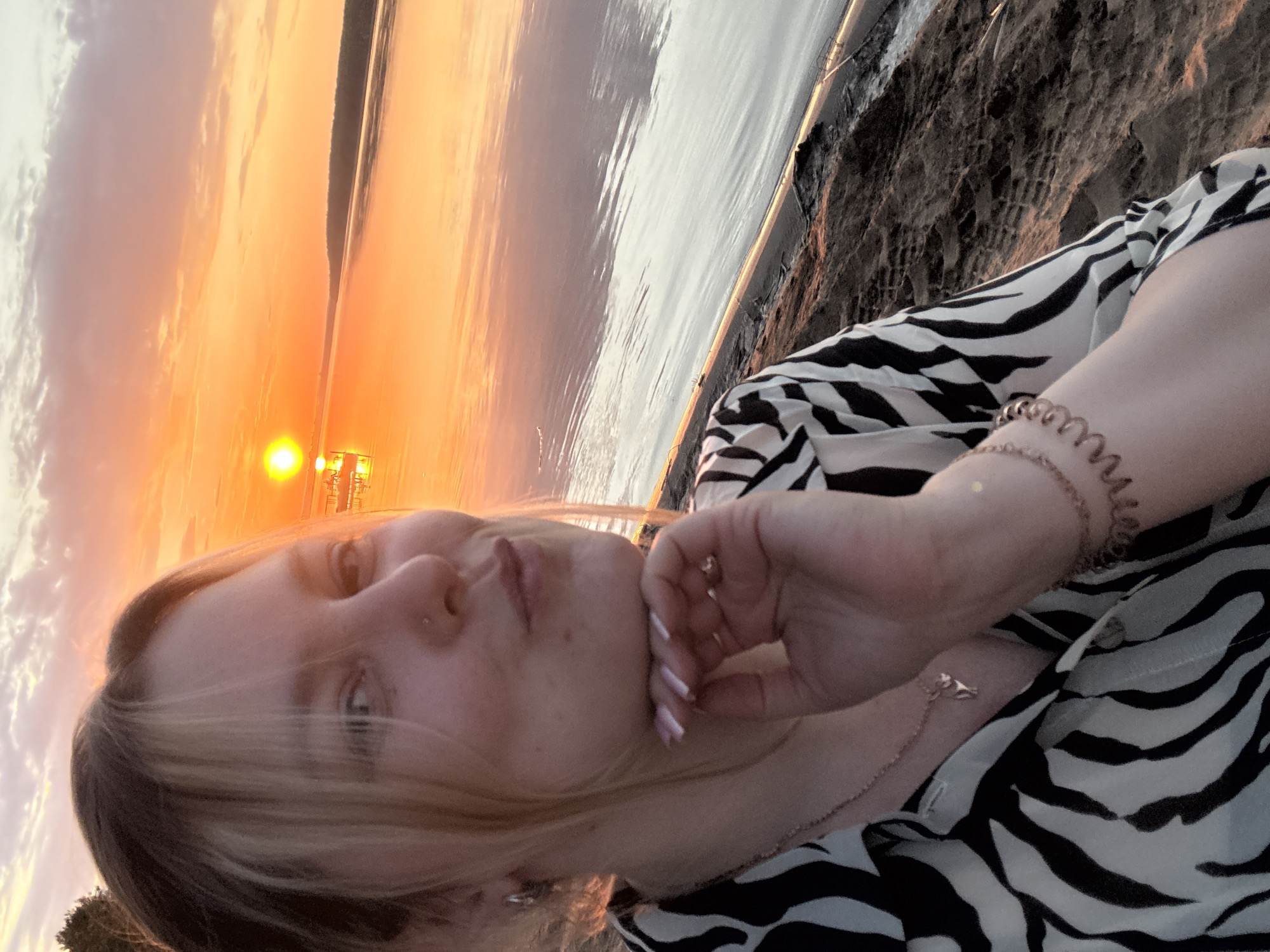 A young person with blonde hair poses for a selfie at sunset. The background shows a beach, water, and sky with a vibrant orange and yellow sunset. In the distance, some industrial structures are visible. The person wears a black and white zebra-striped shirt and a bracelet. The location is likely near Chaykovsky, Russia, given the prompt context, but the specific location within the city is unclear from this image.