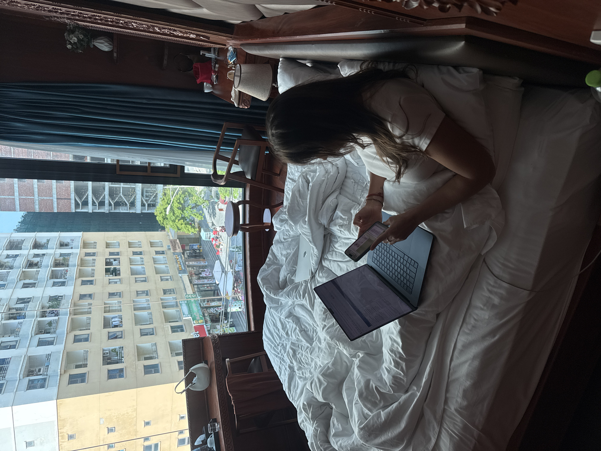 A person sits in a bed covered in white bedding with a laptop on the bed and a mobile phone in their hand. The person has long brown hair and is wearing a white shirt. A large window behind the person looks out onto a city street in Đà Nẵng, Vietnam. Buildings are visible outside the window. A side table with a lamp sits next to the bed.