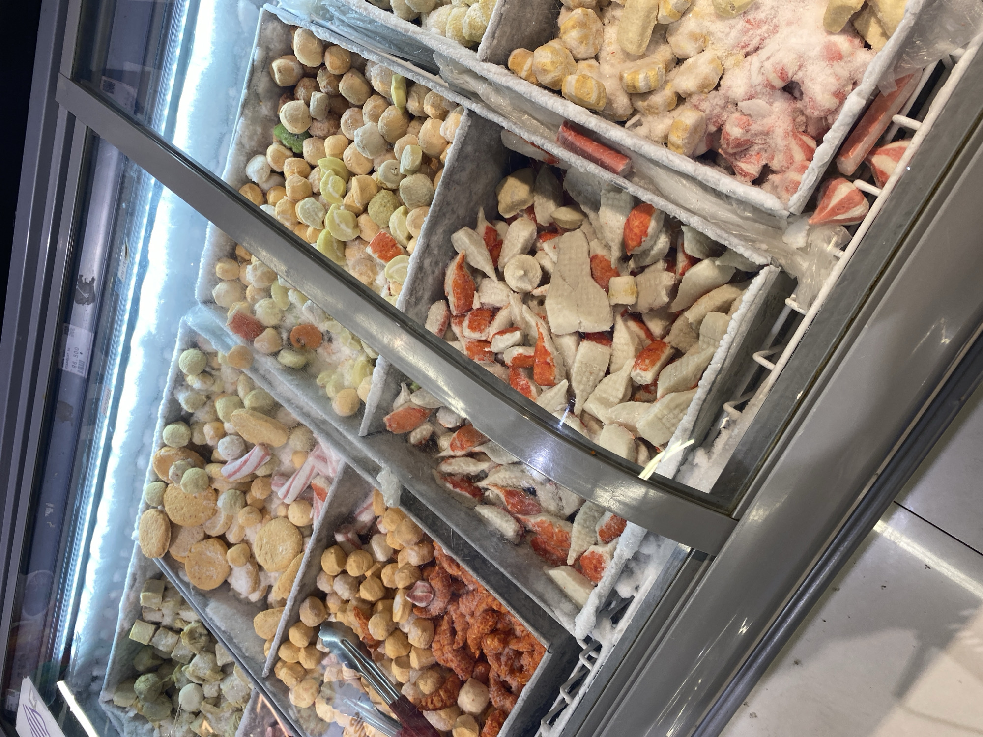 This image shows a freezer display case in a market in Banda Aceh, Indonesia, filled with various types of frozen seafood products. The display is divided into multiple compartments, each containing a different item. From left to right, the compartments appear to hold:
* **Top left:** Cubed, light-colored frozen items, possibly fish cakes or tofu.
* **Second from top left:** Round, golden-brown items, likely fish balls or similar processed seafood. Mixed in are some flat, circular items and a few red and white striped pieces, resembling imitation crab sticks.
* **Third from top left:** A compartment filled with bright orange, processed shrimp or seafood. Tongs are visible in this compartment.
* **Fourth from top left:** A large compartment filled with a mix of white and orange pieces of what appears to be imitation crab meat, some of which are shredded or flaky.
* **Top middle:** A variety of round, yellowish, and light green frozen balls, possibly different types of fish or seafood balls. Some red and white striped pieces are also present.
* **Second from top middle:** Similar to the compartment to its left, this one contains numerous round, yellowish and light green balls.
* **Bottom middle:** More imitation crab meat, similar to the compartment to its left, with white and orange pieces.
* **Top right:** A compartment with more round, yellowish balls.
* **Second from top right:** A compartment filled with yellowish balls, some appearing to have a striped pattern.
* **Bottom right:** A compartment containing frozen, processed seafood with a distinctive pink and white striped pattern, possibly imitation crab or fish cakes.
The items are covered in frost, indicating they are kept at a very low temperature. The freezer display has a glass front with a metal frame, and fluorescent lighting illuminates the products from above. The floor beneath the display appears to be tiled. The overall scene depicts a typical retail environment for frozen foods in Indonesia.