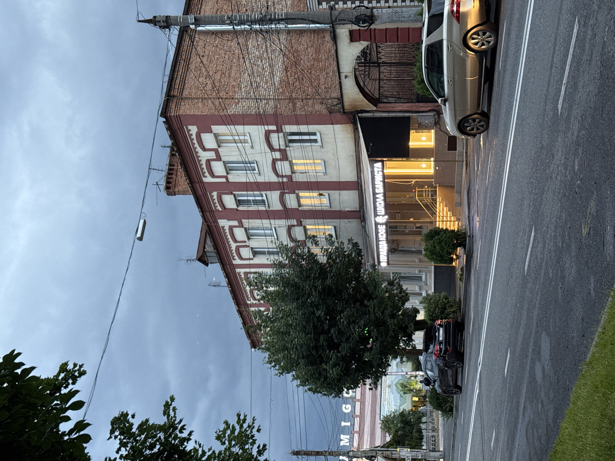 The image depicts a street scene in Assinovskaya, Russia, on an overcast day. A red and white brick building with multiple windows dominates the right side of the frame. The building has a sign above its entrance that reads "ПРАВДА КОФЕ ШКОЛА БАРИСТА" (Pravda Coffee Barista School). In front of the building, there is a modern entrance with yellow lighting.
To the left of this building, a large green tree stands prominently. Further to the left, a pink and white structure with the partial text "M I G" is visible in the background. A black SUV is parked on the side of the road in front of the tree and the pink building. A light-colored sedan is parked on the road to the right of the "Pravda Coffee" building, its rear illuminated by the setting sun. The street in the foreground is asphalt with white lane markings. A grassy area is visible in the lower left corner. Overhead, electrical wires crisscross the sky, and a lamppost is present on the left side of the road. The sky is cloudy and muted in color.
