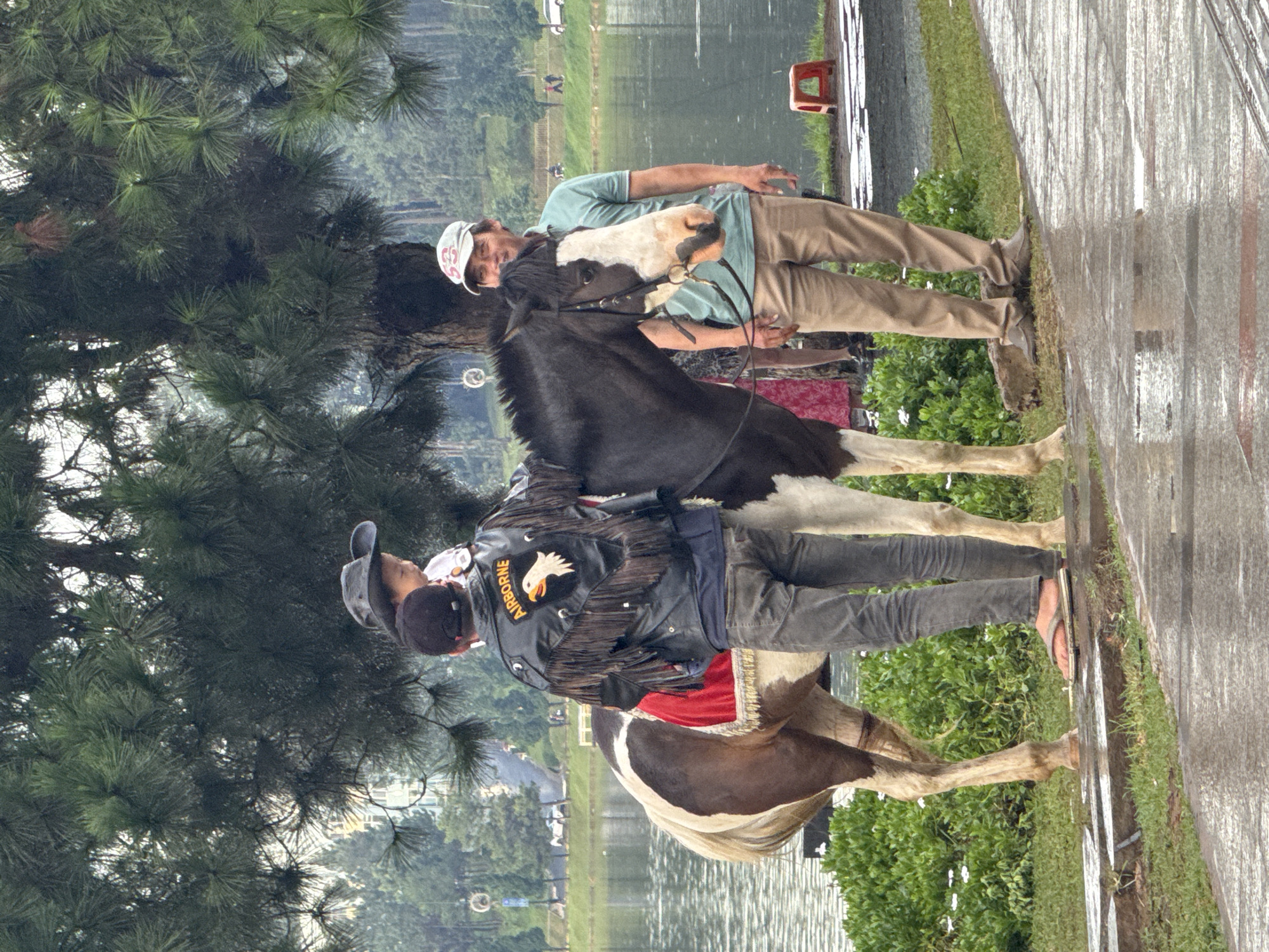The image depicts a scene near Xuan Huong Lake in Da Lat, Vietnam. In the foreground, a man wearing a black leather jacket with fringe and the word "AIRBORNE" and an eagle emblem on the back stands next to a piebald horse. A young child, wearing a cowboy hat, is being held by the man, sitting on the horse. The horse has a red saddle blanket with gold trim. To the right of the horse and man, another man stands looking at the camera, wearing a light blue polo shirt, khaki pants, and a baseball cap. He appears to be associated with the horse and rider. The background shows the lake, lush green trees, and distant buildings, characteristic of Da Lat's landscape. The ground in the foreground appears wet, suggesting recent rain.