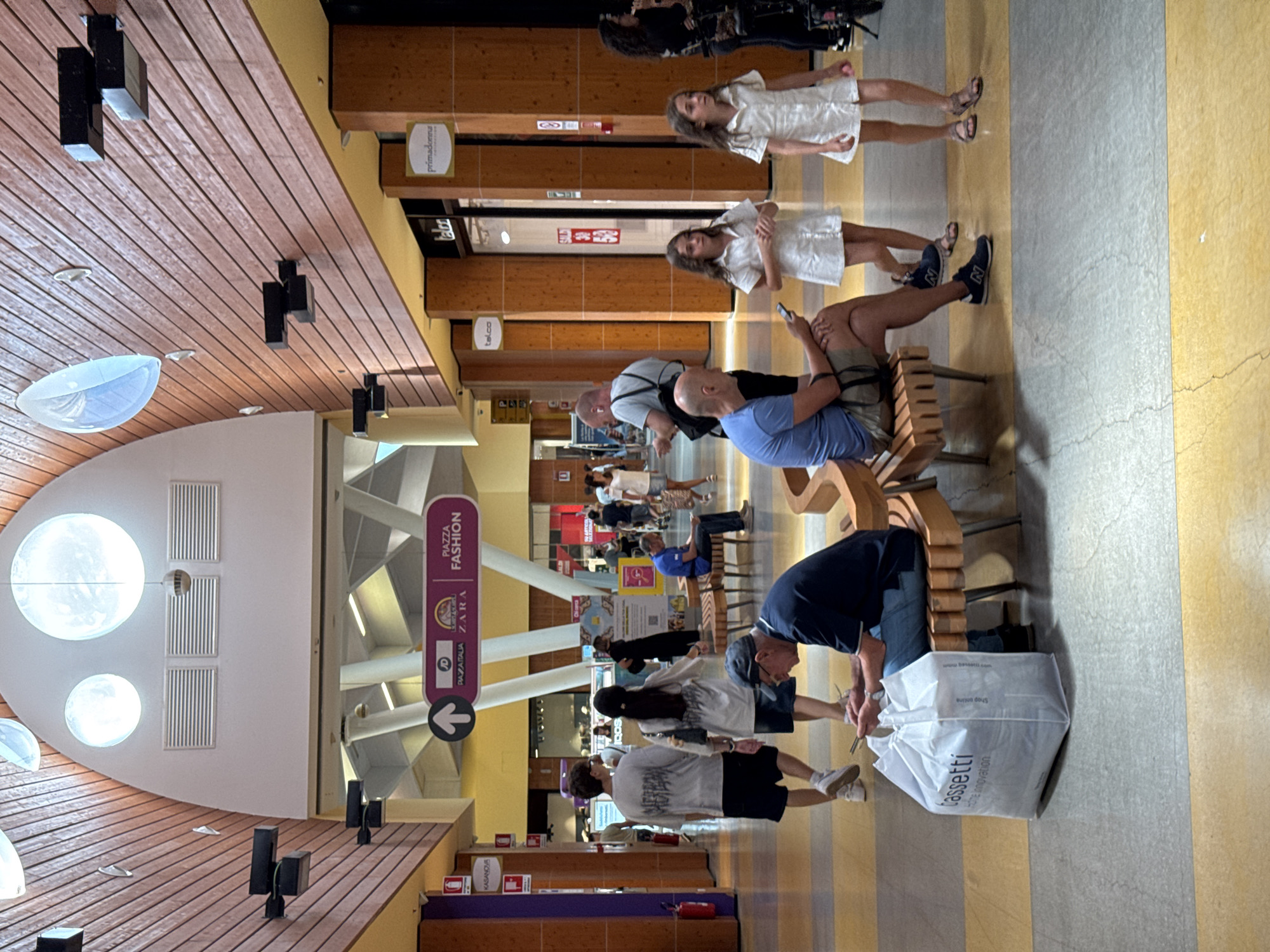 This image depicts the interior of the Forum Palermo shopping mall, located in Palermo, Italy. The scene captures a busy shopping environment with various individuals present. In the foreground, a man wearing a blue t-shirt and shorts is seated on a wooden bench, looking at his phone. Beside him, on the floor, is a large white shopping bag with the "cassetti" logo. To his right, two young girls in white dresses are walking, one looking towards the camera. Further in the background, other shoppers are seen walking and browsing. The architecture of the mall is visible, featuring a wooden-paneled ceiling with circular light fixtures and a wide walkway with yellow and grey floor tiling. Signage for various stores, including "JD," "ZARA," and "PIAZZA FASHION," is displayed. The overall impression is a vibrant and active commercial space.