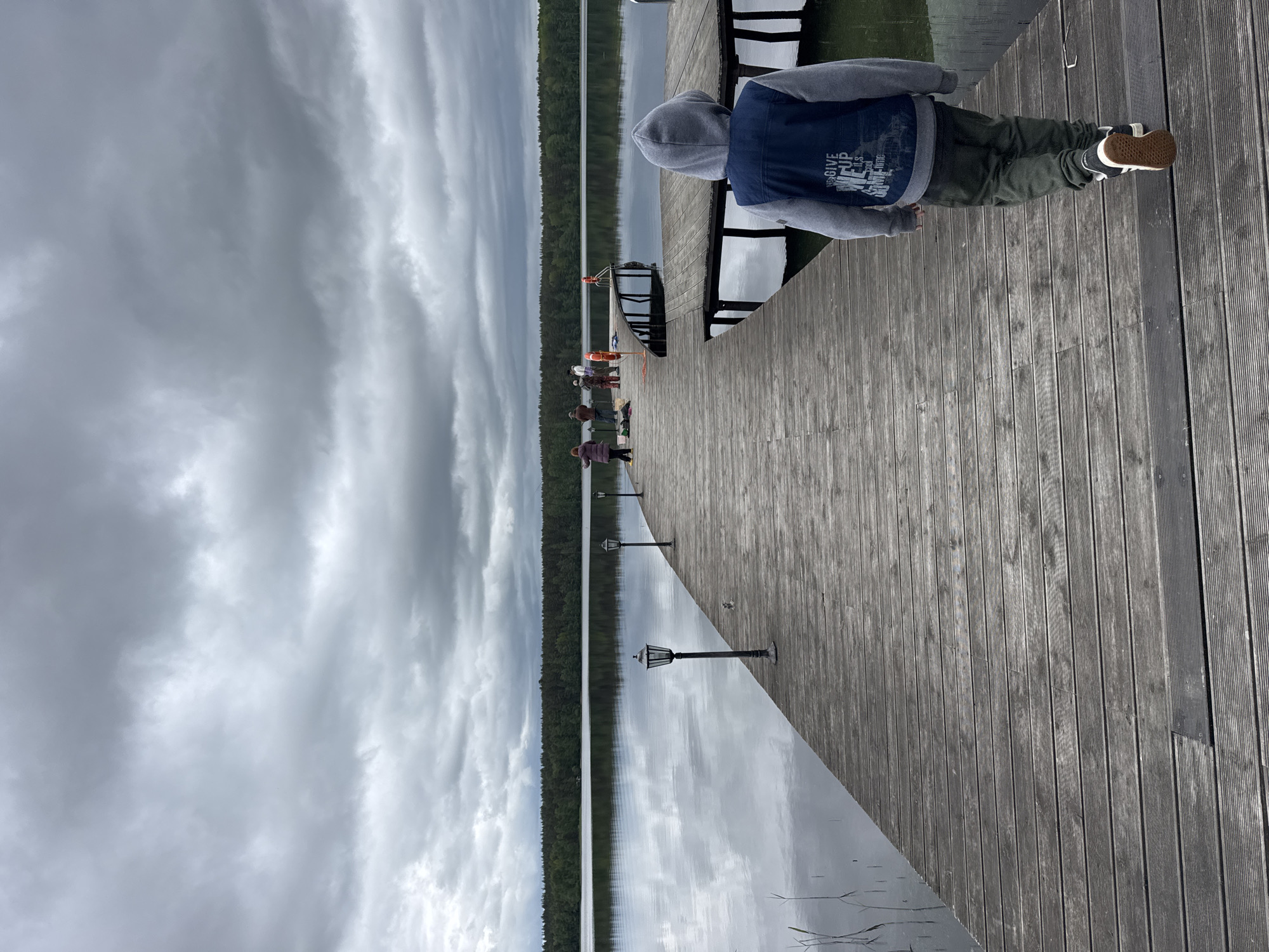 A child in a hooded sweatshirt, dark pants, and sneakers stands on a wooden dock, looking out towards a lake. Other people stand further down the dock, near boats. The sky is cloudy. The location is likely Aleksandrovka, Russia.