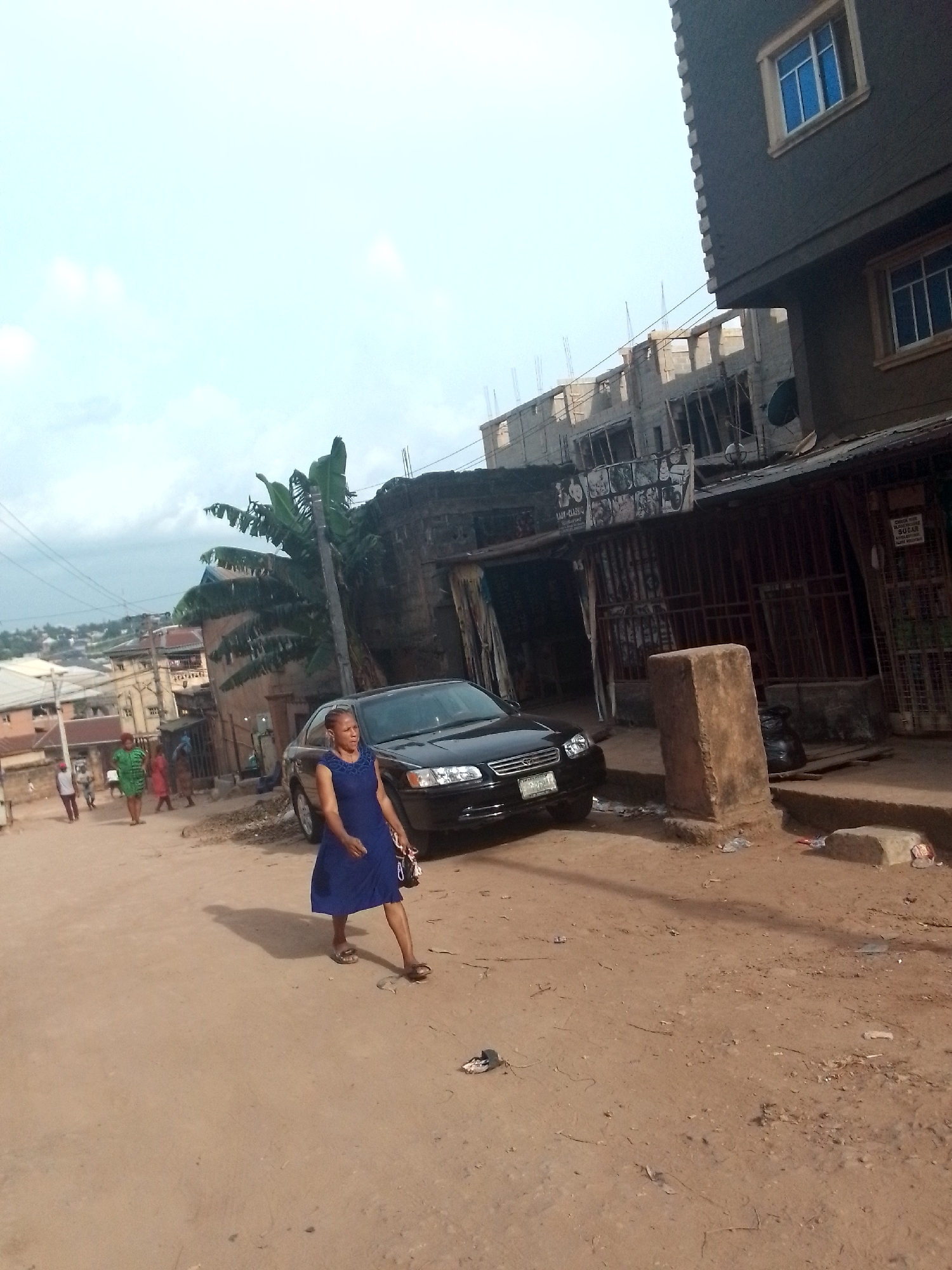 The image depicts a street scene in Onitsha, Nigeria. The street is unpaved, appearing as dirt or reddish soil. A woman in a blue dress walks along the street. A black car is parked near a building. Several individuals are walking in the background. The architecture is mixed, with both older and newer buildings visible. A banana tree is located near the road. The sky is overcast.