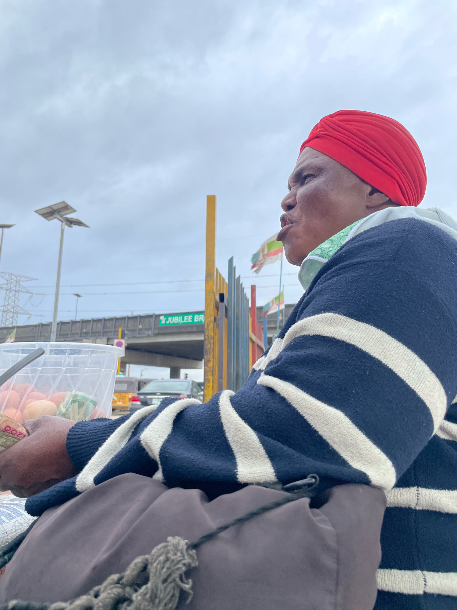 An adult person wearing a red headscarf and a navy blue and white striped sweater is pictured from the side in Ajah, Nigeria. The person is seated, with their upper body visible from the shoulder up. In front of them is a clear plastic container holding a light brown, round objects, presumably eggs, and some folded currency. A gray bag and some grey rope are in the foreground. In the background, a yellow pole stands next to a fence with vertical bars in various colors. Above the fence, a highway overpass is visible with a green sign that reads "JUBILEE BR". Solar-powered streetlights are positioned on tall poles. The sky is overcast and cloudy.