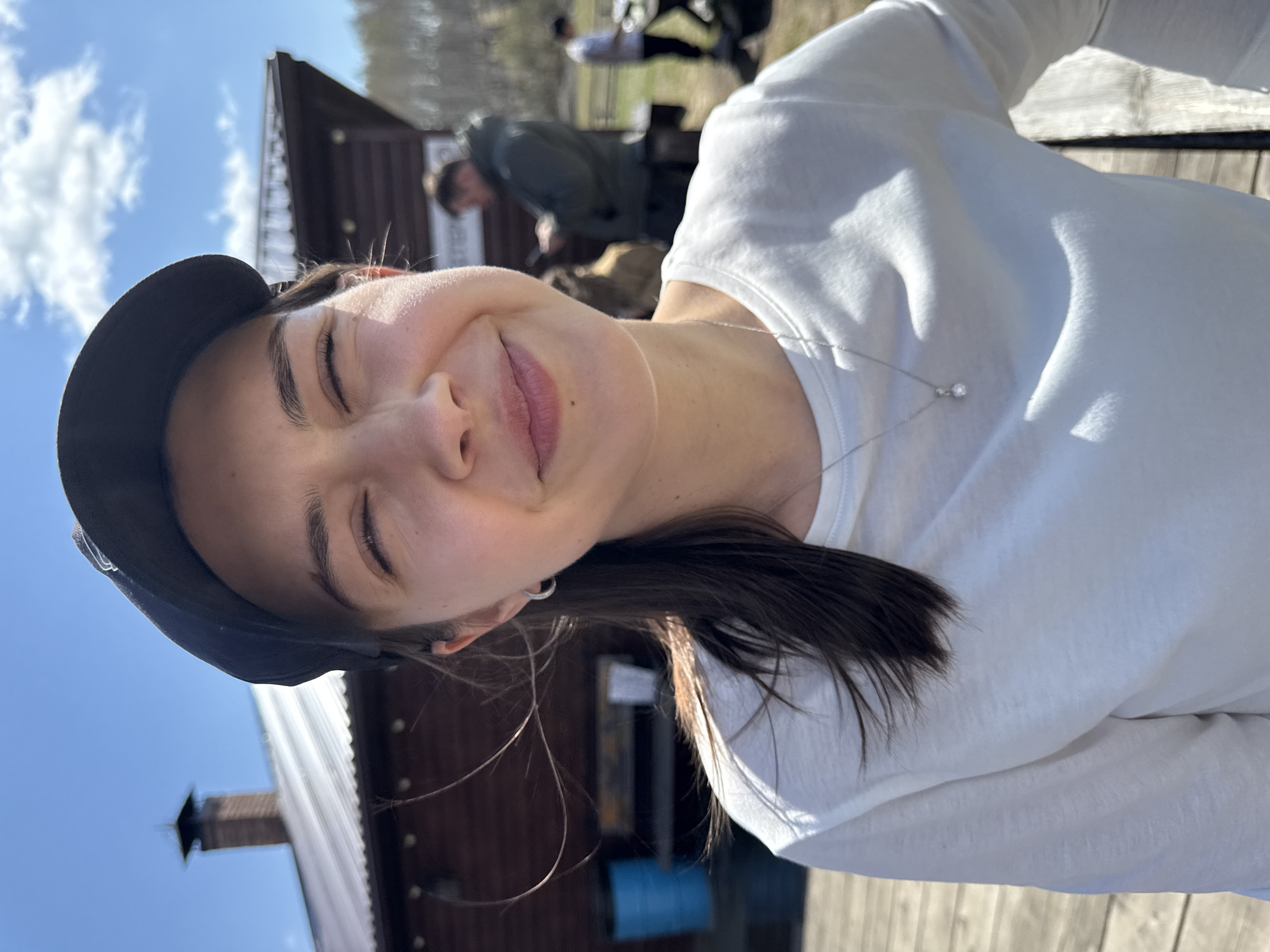 A young woman is posing for a photo outdoors, near Старцево, Russia. She is wearing a dark cap and a white long-sleeved shirt, and she has a silver necklace. Her eyes are closed, and she is smiling slightly. In the background, there is a wooden building with a chimney, a blue barrel, and people moving around. The sky is blue with some clouds. The ground appears to be covered in wood chips.