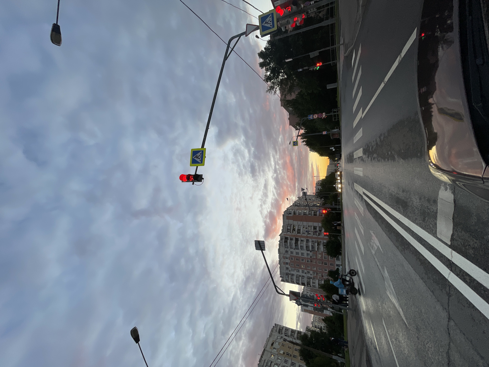 The image depicts a street scene in Moscow, Russia, likely during twilight. The sky is filled with dramatic clouds, tinged with pink and orange from the setting sun. Several traffic lights are visible, with some showing red, indicating that traffic is stopped. A pedestrian crossing is marked on the wet asphalt road, suggesting recent rain.
In the foreground, a portion of a dark-colored car is visible on the right side of the frame. On the road, a person is riding a scooter, wearing a blue jacket. Other people are seen near the sidewalk. Two streetlights are also present in the scene. Tall apartment buildings line the street, characteristic of urban Russian architecture.
The overall mood is calm and transitional, reflecting the end of the day. The wet road and cloudy sky suggest damp weather. There are no visible text elements in the image.