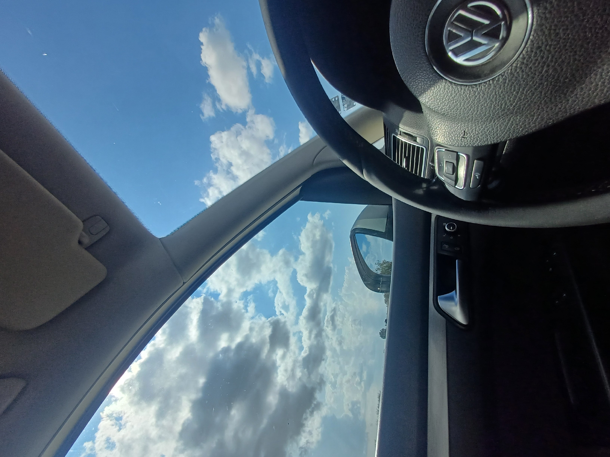 The image is taken from inside a car, looking upwards towards the sky and outwards through the windshield and side window. The subjects include parts of the car's interior: the steering wheel with a Volkswagen logo, part of the dashboard with air vents and buttons, a sun visor, a door handle, and the interior of the car door. The exterior scene visible through the windows is a bright blue sky filled with white, fluffy clouds. The overall setting is a car, likely on a road, given the view of the sky and the presence of a side mirror.
The activity taking place is passive observation of the sky from within the vehicle. It is daytime, indicated by the bright sky and clouds. The weather appears to be clear with scattered clouds, suggesting a pleasant day. There are no people visible in the image. The lighting suggests it could be mid-morning or afternoon. No specific location cues beyond it being Bulgaria are provided, but the car interior implies a modern vehicle. There is no discernible emotion conveyed, as the image is focused on the environment and car interior.
Visible text includes the Volkswagen logo on the steering wheel. There is also some small, illegible text on a sticker on the steering column, and possibly on the buttons on the steering wheel. The overall impression is one of a serene moment, with the driver or passenger enjoying the view of the sky from the comfort of their car.
