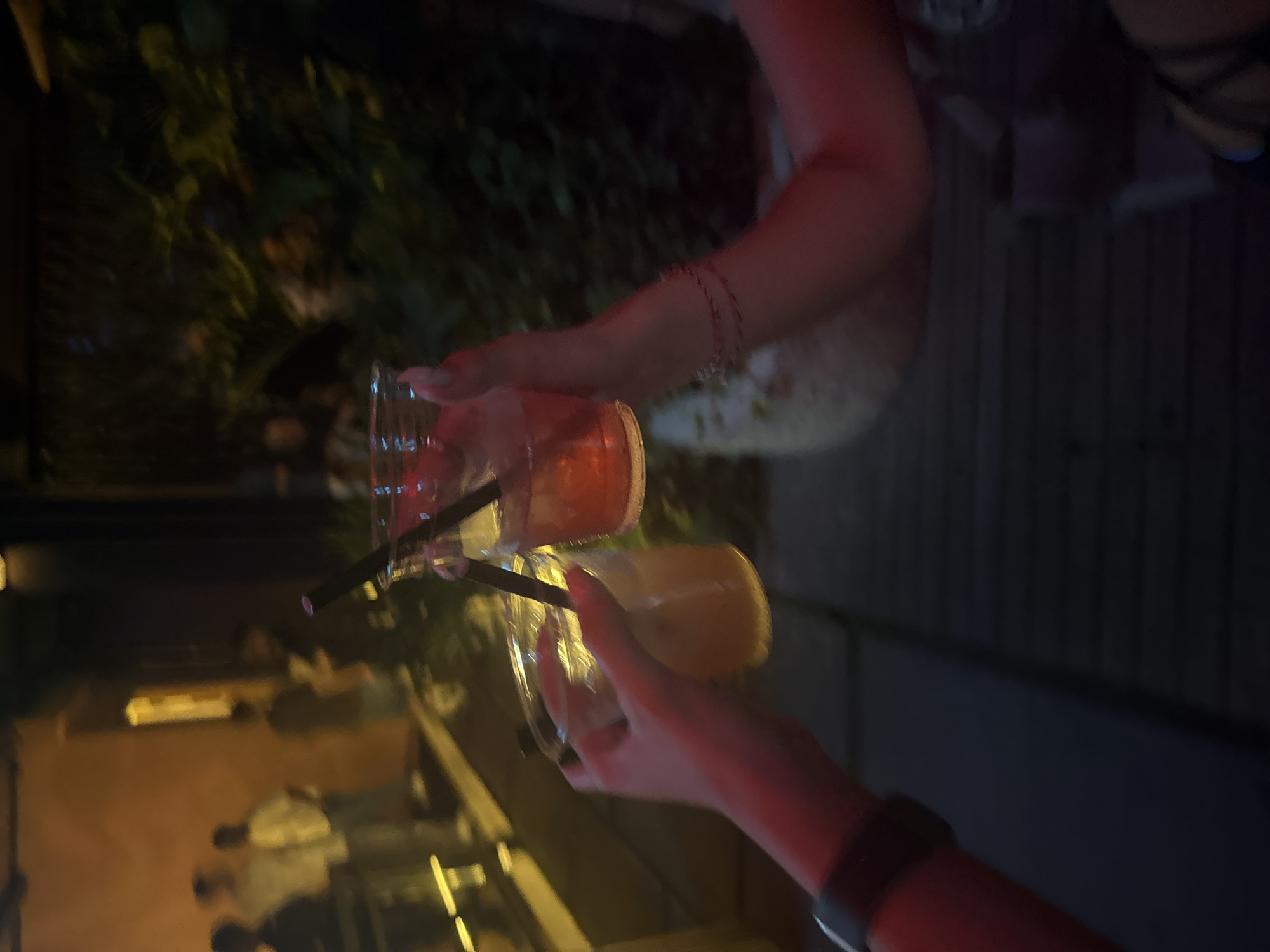 An indoor or semi-outdoor night scene in North Kuta, Indonesia, captures two individuals raising their drinks in a toast. The image shows two hands holding clear plastic cups, appearing to clink them together or hold them up in a gesture of celebration.
On the left side of the frame, one hand holds a cup filled with a vibrant orange-red beverage, containing a black straw. The arm is visible, adorned with a delicate, multi-strand bracelet. On the right, another hand holds a similar clear cup containing a lighter, yellowish or beige drink, also with a black straw. This individual wears a black smartwatch or band on their wrist. Both individuals' arms are illuminated with a warm, reddish glow, suggesting ambient lighting from the venue.
The background is dark and largely out of focus, featuring blurry green foliage, indicative of plants or bushes, and faint, warm artificial lights in the distance. Other indistinct figures of people are dimly visible further back, suggesting a social gathering at a bar or restaurant setting. The activity conveys a sense of celebration or camaraderie among friends. No text is visible in the image. The time of day is clearly nighttime or late evening due to the darkness and reliance on artificial lighting.
