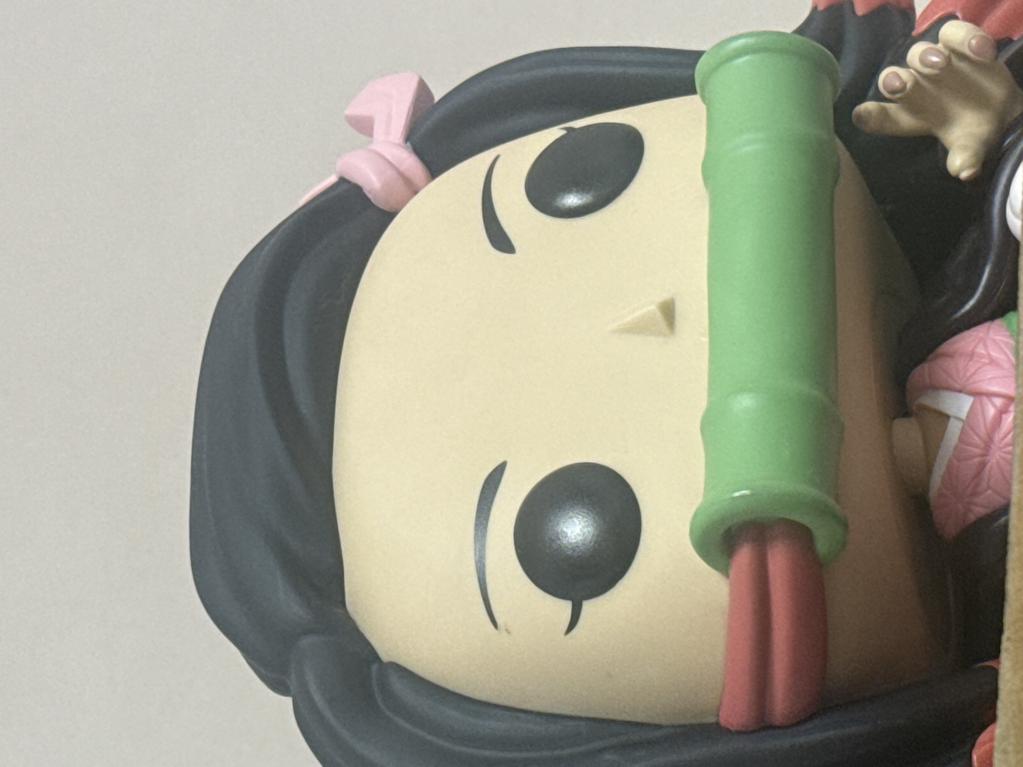 The image is a close-up, head-on shot of a Funko Pop figure. The figure has black hair, a pale complexion, and large black eyes. It is wearing a pink hair accessory on the right side of its head and a green bamboo-like muzzle that covers its mouth. The figure is wearing a pink garment with a geometric pattern. A portion of its right hand is visible, with fingers extended and painted fingernails. The background is a plain, light-colored wall. The figure is positioned against a light-colored surface, possibly a shelf or table. No specific location in Nha Trang, Vietnam can be determined from the image.