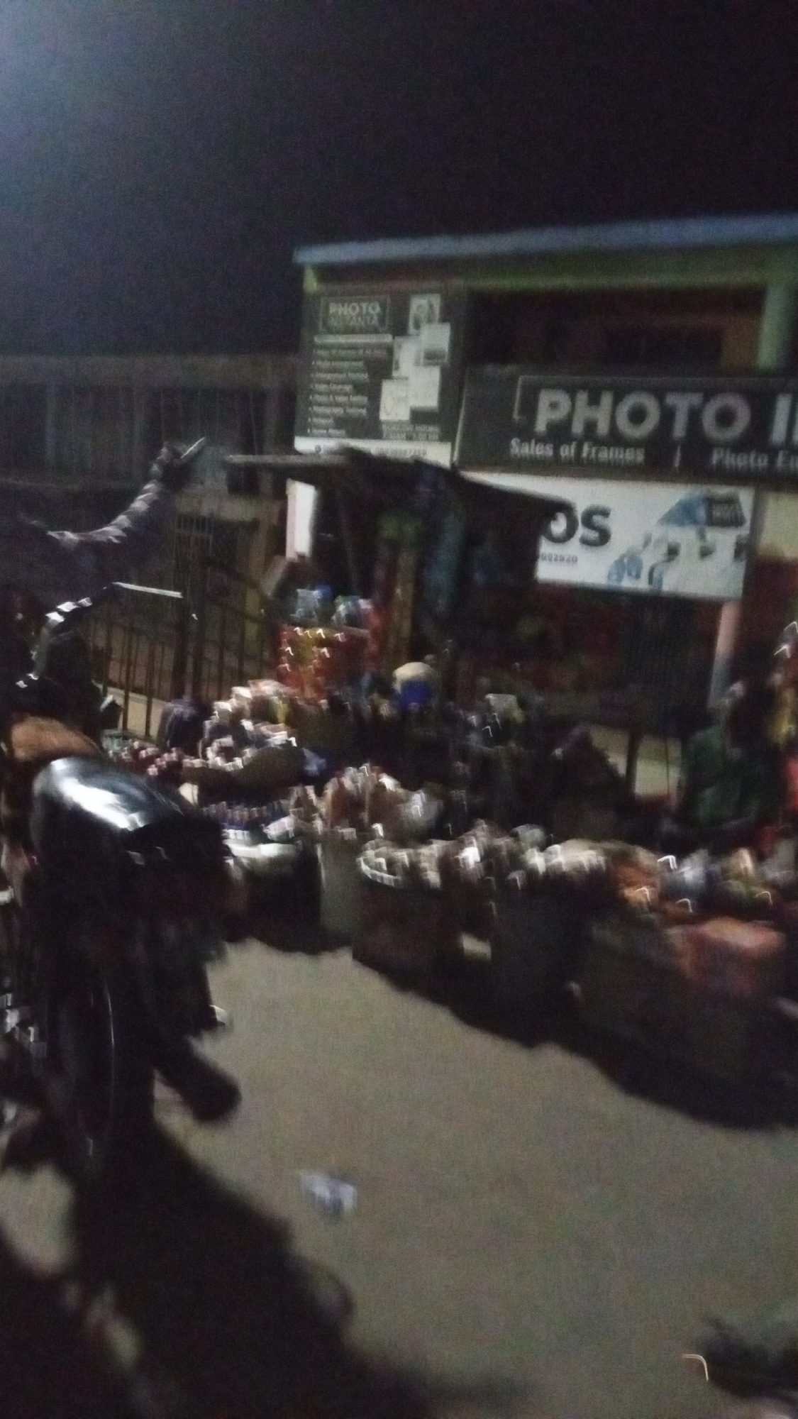 This blurry and dark image captures a street scene at night, likely in Lokoja, Nigeria. The setting appears to be an active market or vending area, indicated by numerous baskets and sacks filled with various goods, possibly produce or food items, spread out on the ground in the middle and foreground.
On the left side of the frame, a dark motorcycle or scooter is partially visible. Indistinct figures, likely vendors and customers, are vaguely discernible among the market stalls; one individual wearing green clothing can be seen on the far right.
In the background, a building houses commercial establishments, identifiable by illuminated signs. A prominent sign on the left displays "PHOTO INSTANT" at the top, followed by a list of services, including "Passport Photos," "ID Cards," "Scanning," "Laminating," "Photo Enlargement," "Photo Books," "Digital Camera Repair," and "Photo Editing." A partial phone number "080398722..." is also visible. Another sign on the right side of the building clearly states "PHOTO I..." and "Sales of Frames," with additional fragmented text like "Photo Enl..." and "DS." The overall blurriness suggests either camera shake or motion blur in the low-light conditions.