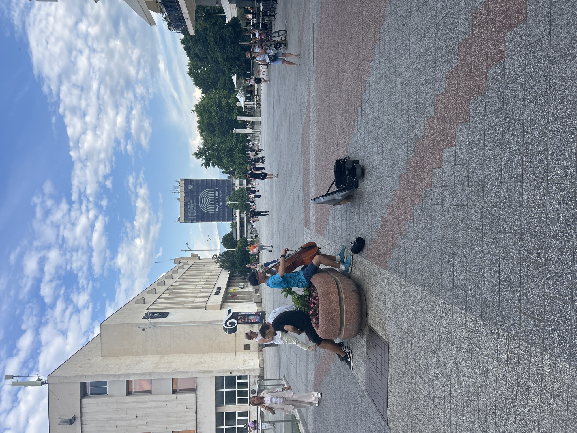 The image captures a vibrant daytime scene in a large, paved pedestrian square in Plovdiv, Bulgaria. In the foreground, two young men are performing music. One, wearing a blue t-shirt and shorts, is seated on a large, oval-shaped planter filled with flowers, playing a cello. Another young man, in a black t-shirt and shorts, stands beside him, possibly also playing an instrument, though it's less clear. An open instrument case, likely for collecting donations, rests on the ground nearby.
The square is paved with patterned grey and reddish-brown granite blocks. In the mid-ground and background, numerous pedestrians are visible, strolling through the public space. To the left, a woman in light-colored clothing walks towards the foreground. On the far side of the square, a modern-looking building stands, and further back, lush green trees line the area.
A prominent large billboard or sign is visible in the distance, displaying text in both Bulgarian and English: "ГРАД ПЛОВДИВ" (GRAD PLOVDIV), "CITY OF PLOVDIV", and "10 ГОДИНИ" (10 GODINI), "YEARS," along with a circular rainbow-like logo. The sky above is bright blue with scattered white clouds, indicating clear and sunny weather. The overall atmosphere is lively and typical of a bustling city center.