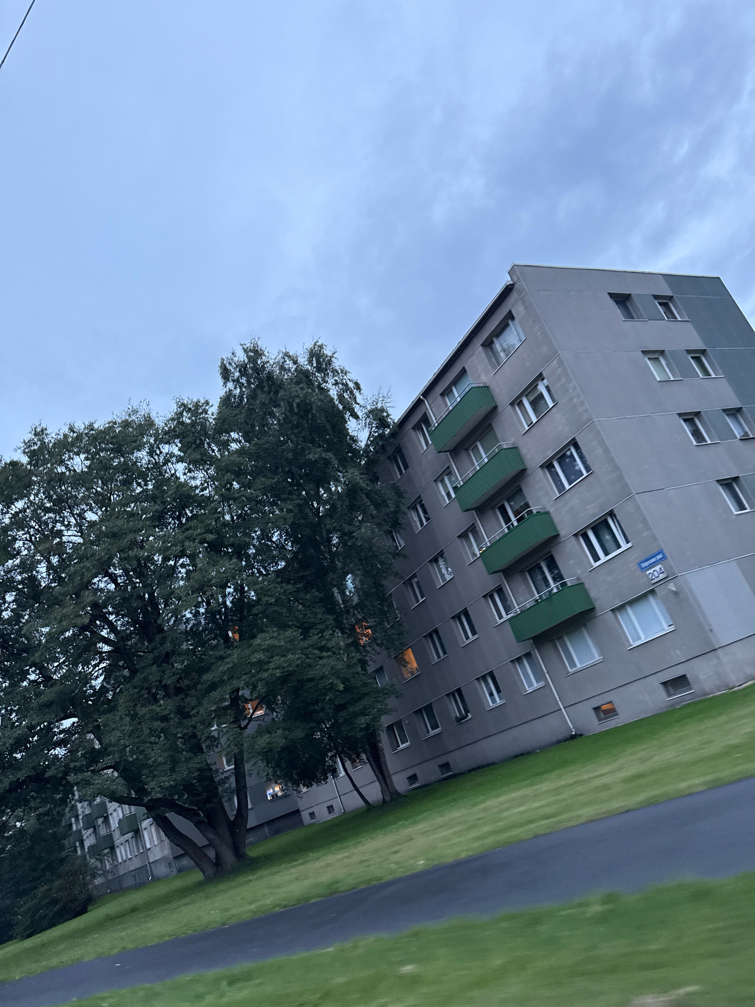 A low-angle, wide shot captures a grey, multi-story apartment building under a cloudy, pale blue sky. The building, with its many windows and green balconies, dominates the right side of the frame. The left side features a large, dark green tree with dense foliage. A grassy slope leads down from the building towards a dark grey asphalt path in the foreground. A street sign with the numbers "204" is visible on the building's facade. The overall scene suggests an urban residential area, likely taken during daylight hours, given the ambient light. The image is tilted slightly, giving a dynamic perspective.