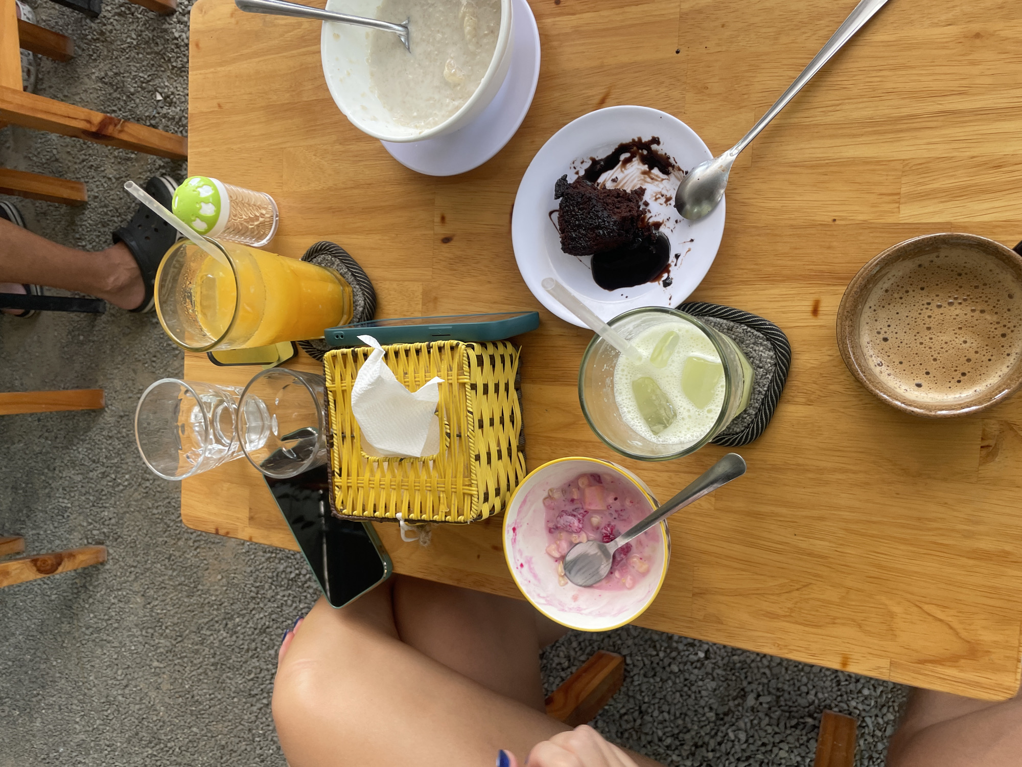 A top-down, medium shot captures a wooden table in an outdoor cafe in Nha Trang, Vietnam, laden with breakfast items and drinks. The table is speckled with gravelly pavement. To the left, a person's bare legs are visible, with a hint of a knee and thigh. On the table, a yellow woven basket with a white tissue dispenser sits prominently. Next to it, a glass of orange juice with a straw is perched on a coaster. Beside the juice, a small container with a green lid, possibly a sugar dispenser, is present. In the center, a bowl of pink yogurt with fruit pieces and a spoon is displayed. To the right of the yogurt, a green beverage with ice and a straw is served in a glass. Further right, a white plate holds a piece of dark chocolate cake drizzled with chocolate sauce and a spoon. A bowl of what appears to be oatmeal or congee with a spoon is positioned in the upper right corner. In the foreground, a ceramic mug filled with coffee sits, showcasing a rich crema. A smartphone lies on the table near the yellow basket. The overall scene depicts a leisurely breakfast or snack break in a relaxed outdoor setting.
