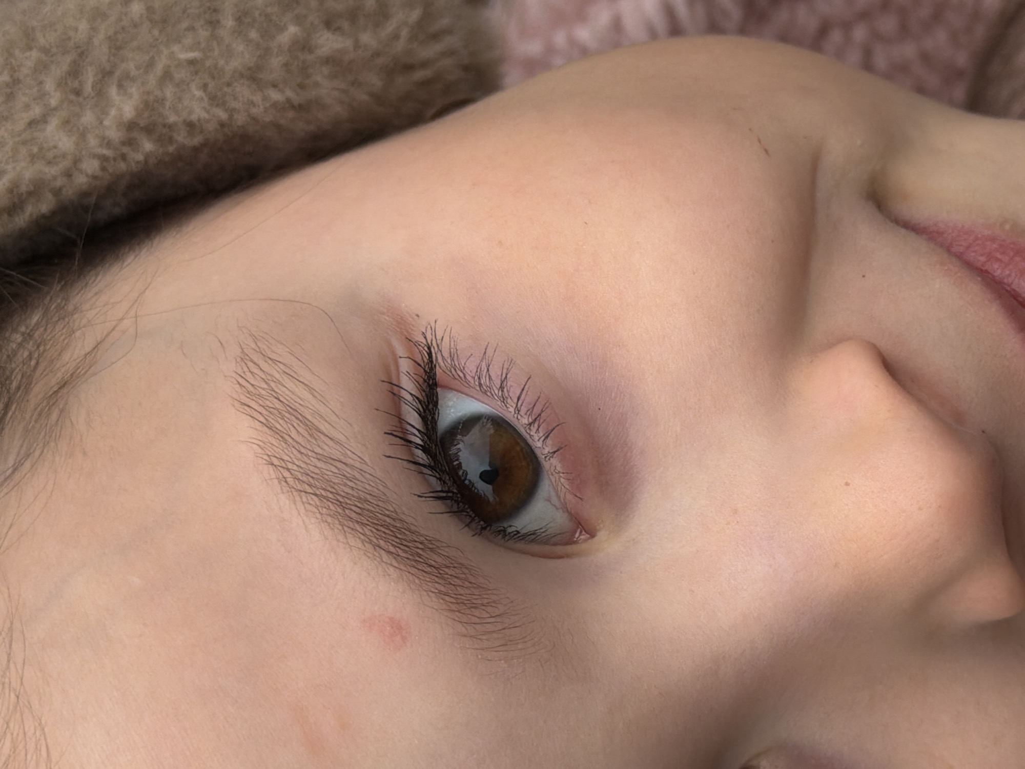 A close-up image shows the face of a young person, likely a child, lying on their side. The focus is on their eye, which is brown with long eyelashes. There is a light brown fuzzy material, possibly a hat or blanket, visible in the upper left corner. The skin is fair. A portion of the nose and lips are visible in the lower right. The overall scene suggests a relaxed or sleeping state. There is no evidence to identify the location in the city of Perm, Russia.