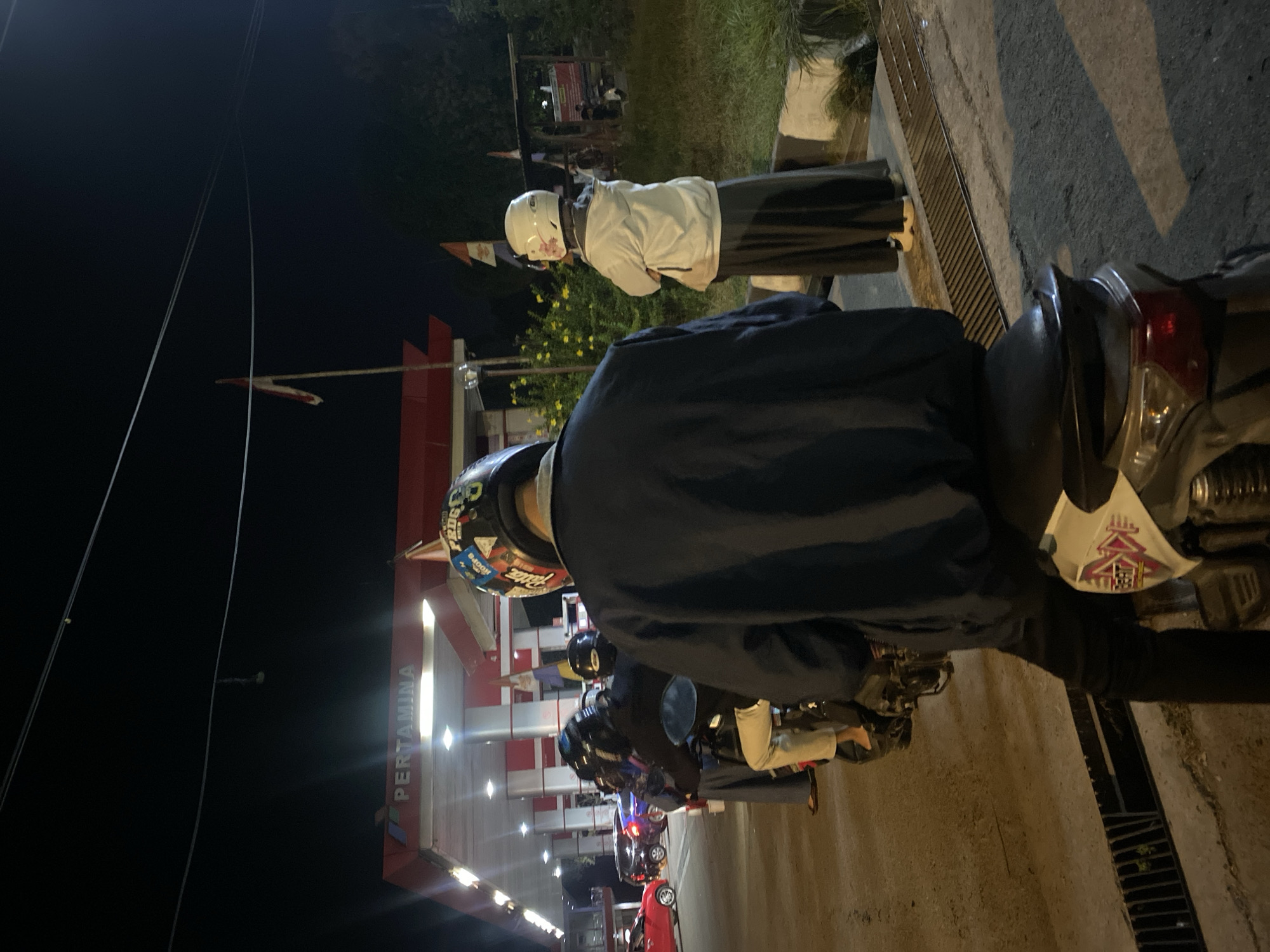 The image captures a nighttime scene at a Pertamina gas station in Gambut Barat, Indonesia. Several individuals are present, primarily on motorcycles. In the foreground, a person wearing a blue jacket and black pants sits on a white and pink motorcycle, their back to the camera. They are wearing a multi-colored helmet with stickers. To the right, a person in a white jacket and dark skirt is standing, also wearing a white helmet with a floral decal. Behind them, a line of motorcycles and people are waiting. In the background, the Pertamina gas station's canopy is illuminated, with fuel pumps visible. Other vehicles, including a red car and a dark-colored car, are also present at the station. The environment appears to be a street or roadside, with pavement, a drainage grate, and some vegetation visible. The overall atmosphere is that of a typical evening at a busy gas station.