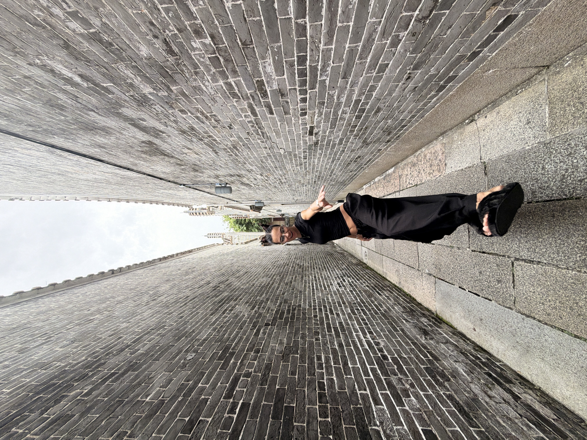 A woman with dark hair pulled up and wearing glasses is standing in a narrow alleyway lined with dark brick walls. She is dressed in black, with a cropped top and wide-leg pants. Her left hand is extended towards the camera, and her right foot is lifted slightly off the ground, as if she is mid-pose. The alleyway is paved with grey stone tiles. The environment suggests an urban setting in Chancheng District, China, characterized by traditional architecture with dark brick construction. The sky above is overcast.